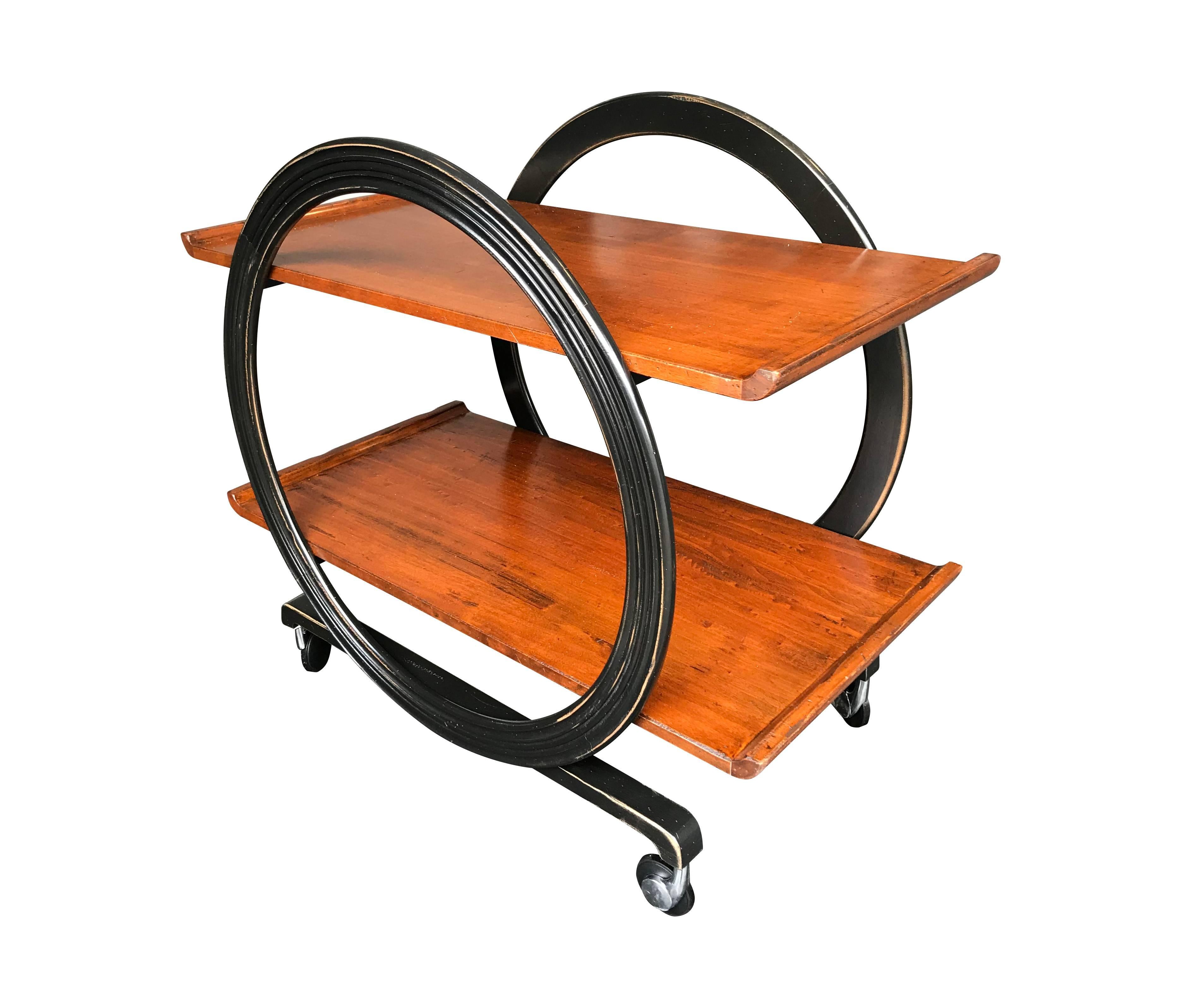 An Art Deco style bar trolley with varnished wooden shelves, with aged finish effect and ebonized circular design frame, on original castors.