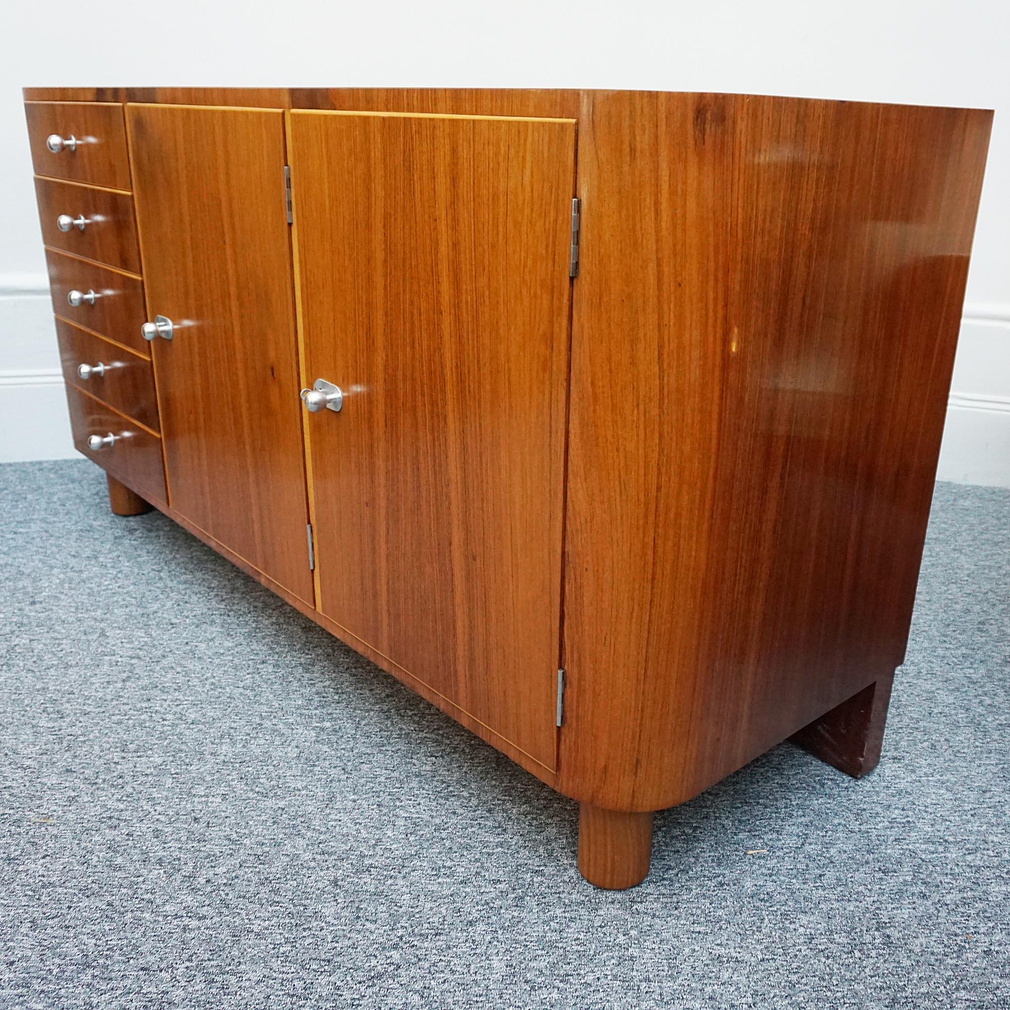 An Art Deco 'Woburn' Sideboard by Gordon Russell. Figured Walnut veneered with elm banding on Solid Oak. Designed in 1933 by Dick Russell. Original chrome handles and keys with five drawers and two shelved cabinet sections. 

Dimensions: H 84.5cm W