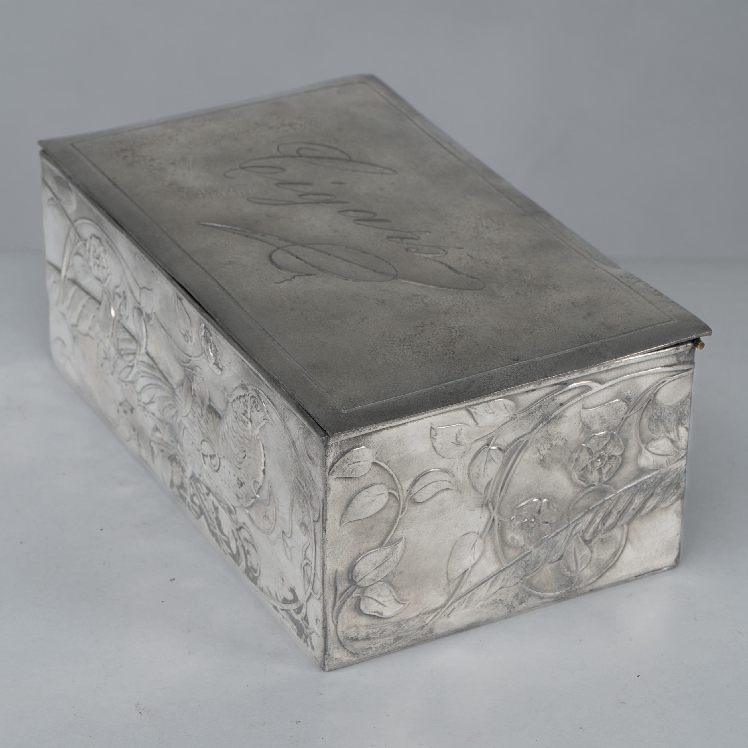An Art Nouveau continental pewter cigar box lined with Spanish cedar and decorated with a pheasant whose tail follows around the side of the cigar box, stood amongst sinuous smokey Nasturtium floral decoration that continues around all four sides.