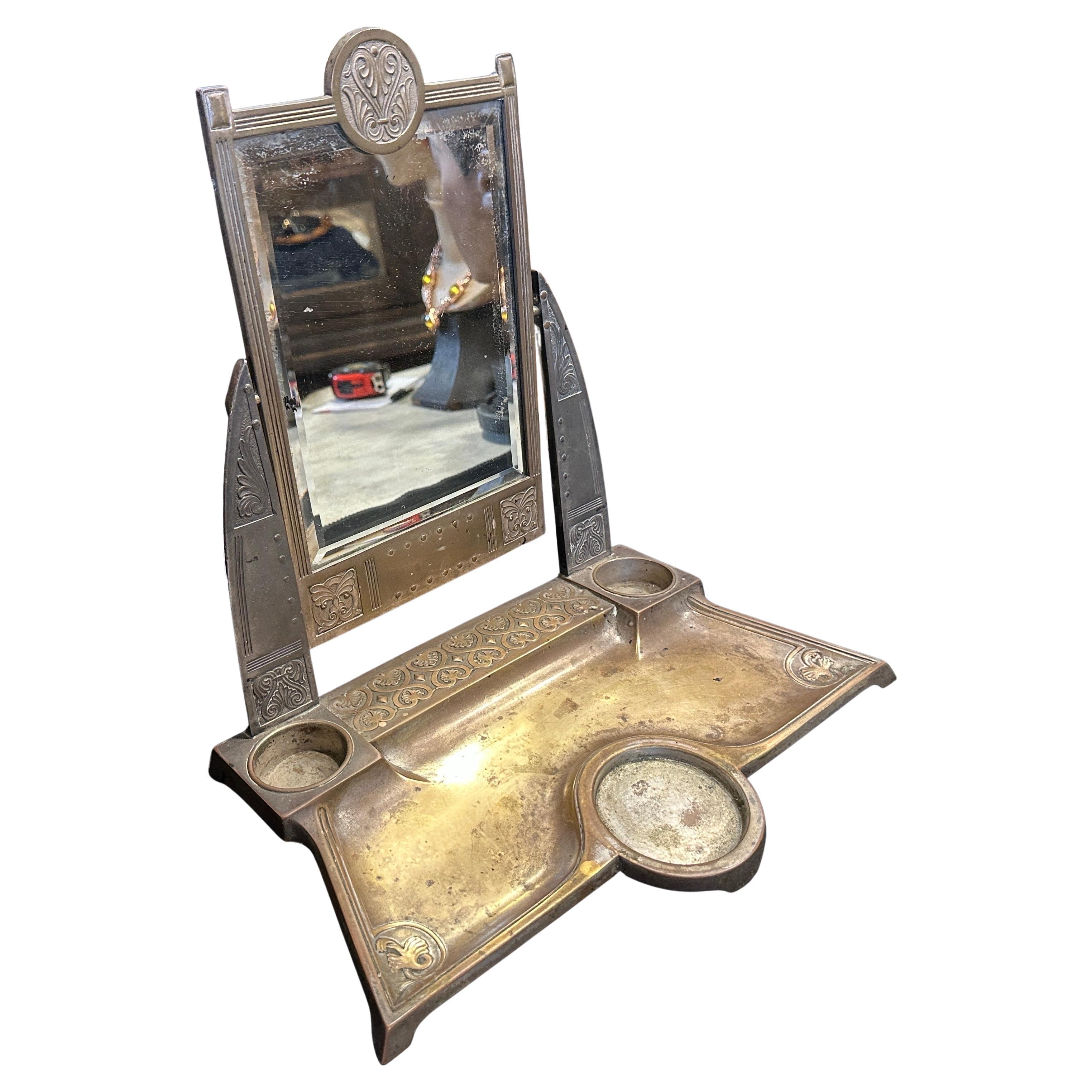 This Art Nouveau table vanity mirror features an elegant, silver-plated design. The mirror is rectangular with a decorative crest at the top, showcasing classic Art Nouveau motifs. It's supported by two ornate side panels, and the base includes