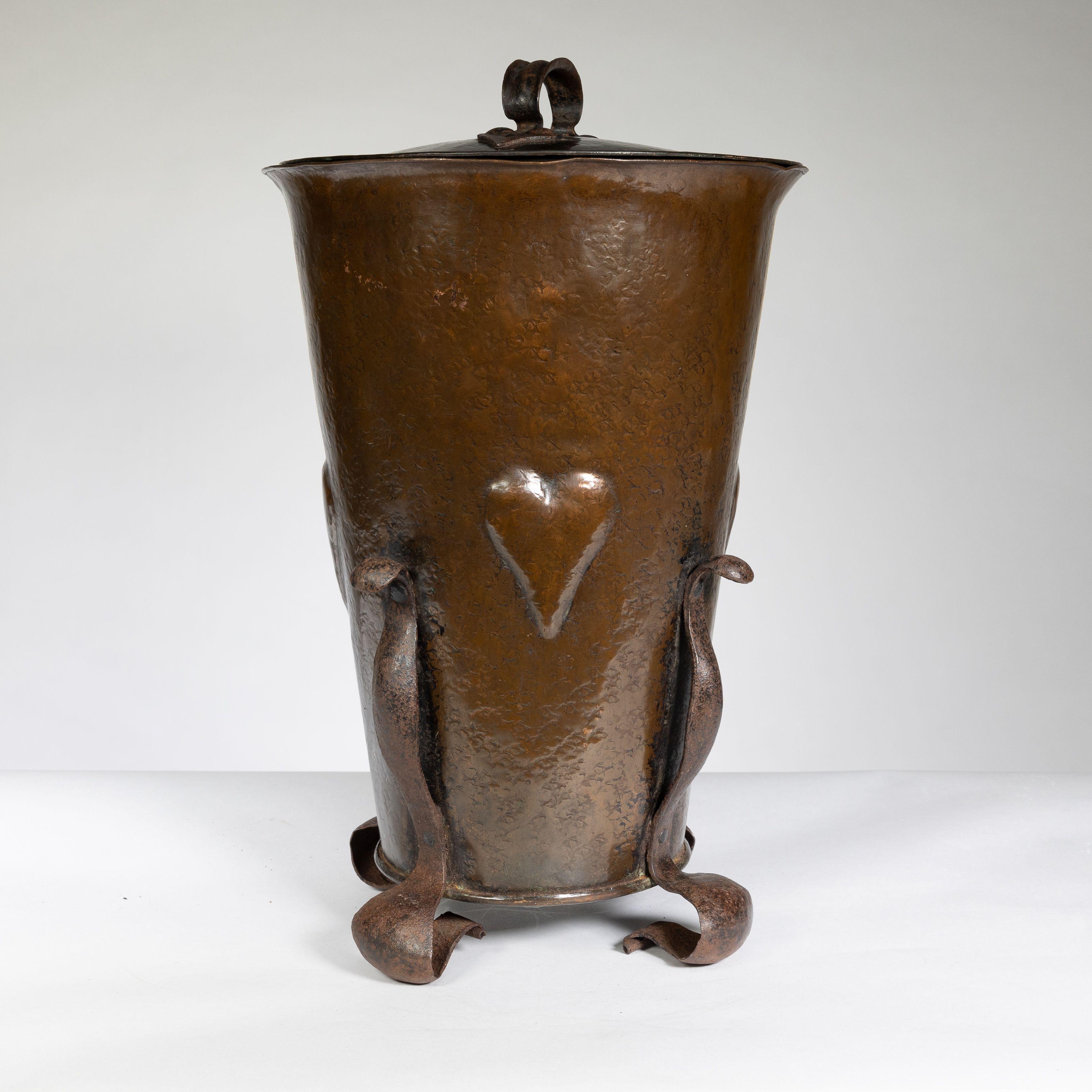 An Arts and Crafts brass coal bucket with a hand wrought iron handle to the lid and four stylized hand repousse hearts with hand wrought iron floral style feet.