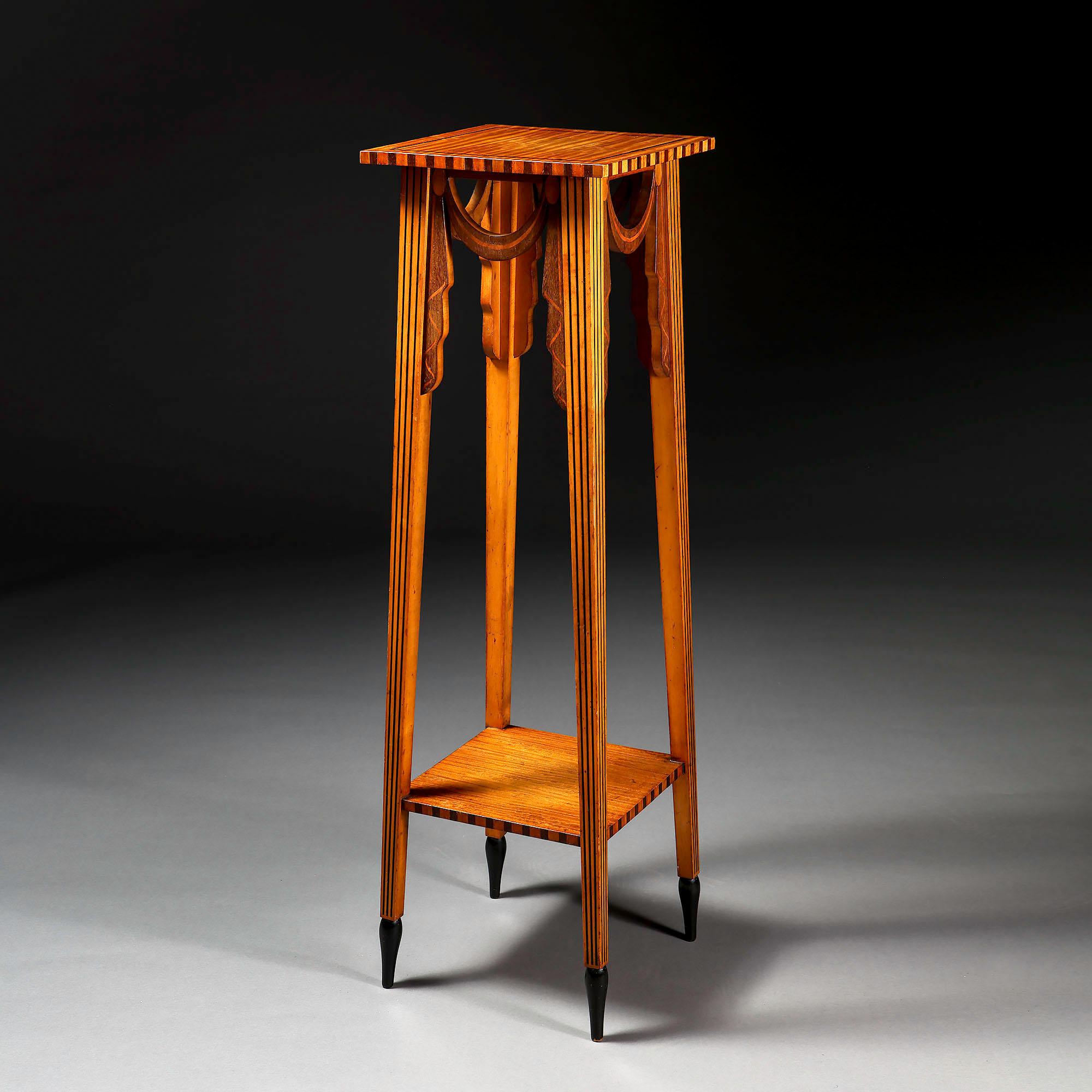An unusual Arts and Crafts satinwood two tier sculpture stand, with swags to the frieze, the edges of the tops with contrasting wood, and the legs with contrasting black wood inlay, and terminating in an ebonised foot.