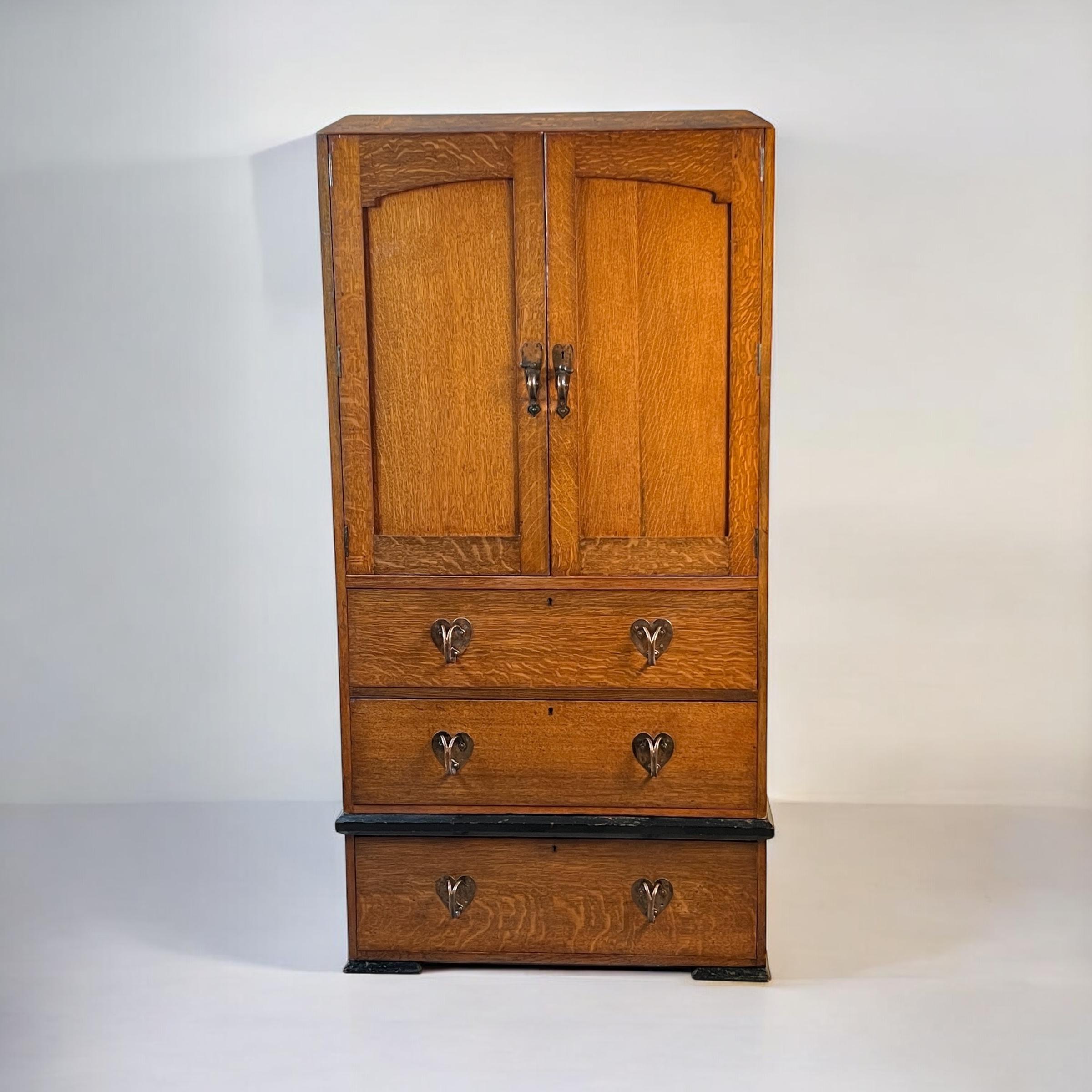 A delightful Arts & Crafts Oak Linen Press in the manner of Liberty London.

It features a subtle, angled cornice at the top, with arched, panelled doors below that enclose three large oak shelves. Below these, sit a bank of three graduated