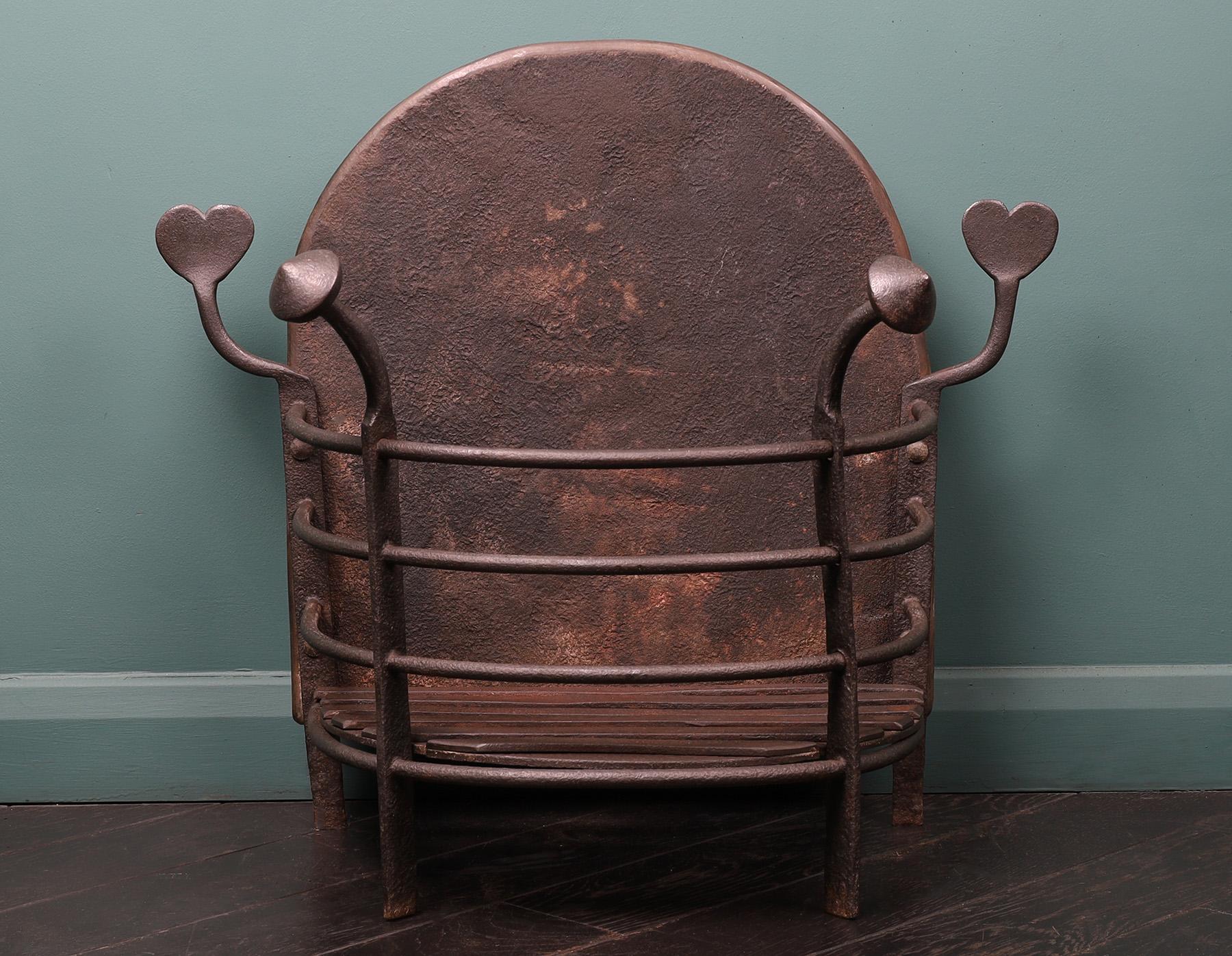 An Arts & Crafts semi-circular wrought-iron fire basket with heart-shaped finials. The railed basket with cone and heart-shaped finials that flank the arched fireback. Wax finish. 
Height of burning area: 4 7/8″
Circa 1880
