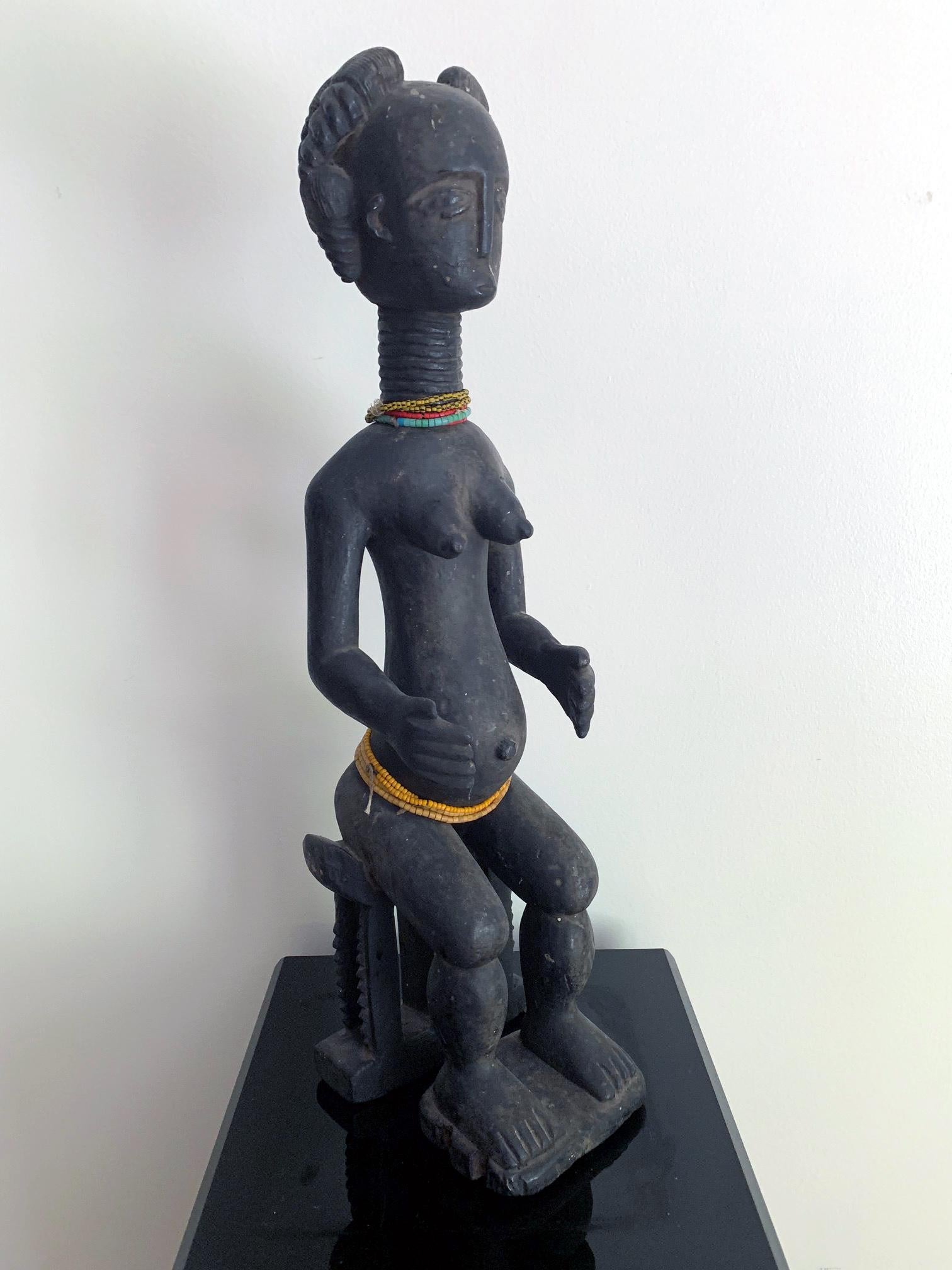 On offer is an Ashanti female fertility figure from Ghana, West Africa, circa Mid-20th century. Carved from a single block of wood, the statue depicts a female with typical coiffure and facial expression on seated on an Akan bench. Her arms