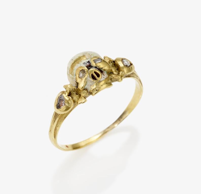 

Memento mori ring-Exceptional antique gold ring with a kitten in the shape of a skull, enamelled in white.
The eyes are set with small rose cut diamonds. The shoulders of the ring are formed by bones.
The body of the ring is enamelled in white and
