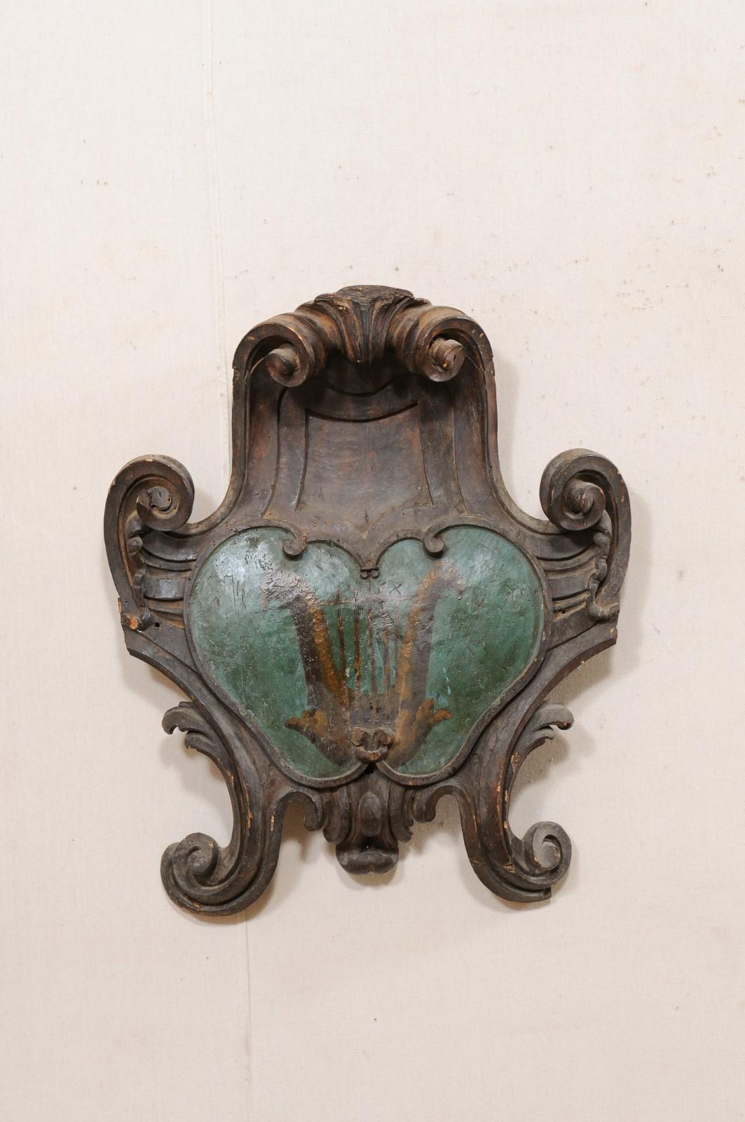 An Italian carved wood family crest wall ornament from the early 18th century. This antique wall plaque from Italy has a beautifully-fluid shape, in hand-carved, thick, scrolls adorning a raised and stylized center, painted with family crest. This