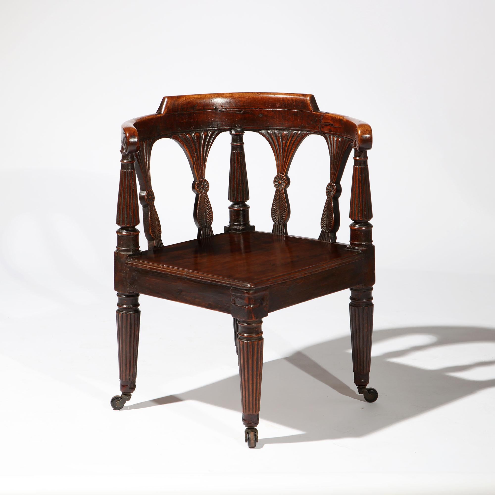 An early 19th century Anglo Indian corner chair in colonial hardwood, with curved back and back splats carved with paterae, with fluted legs terminating in brass castors.