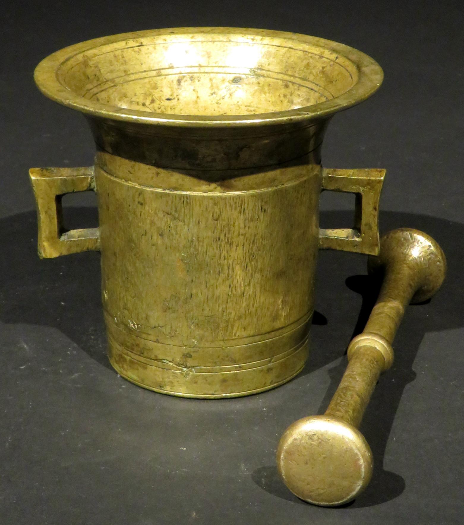 The cast body of cylindrical form rising to a flaring rim, engraved with incised ring turned detail to the exterior, interior and underside, sided by twin open handles together with what appears to be its original pestle with compressed and