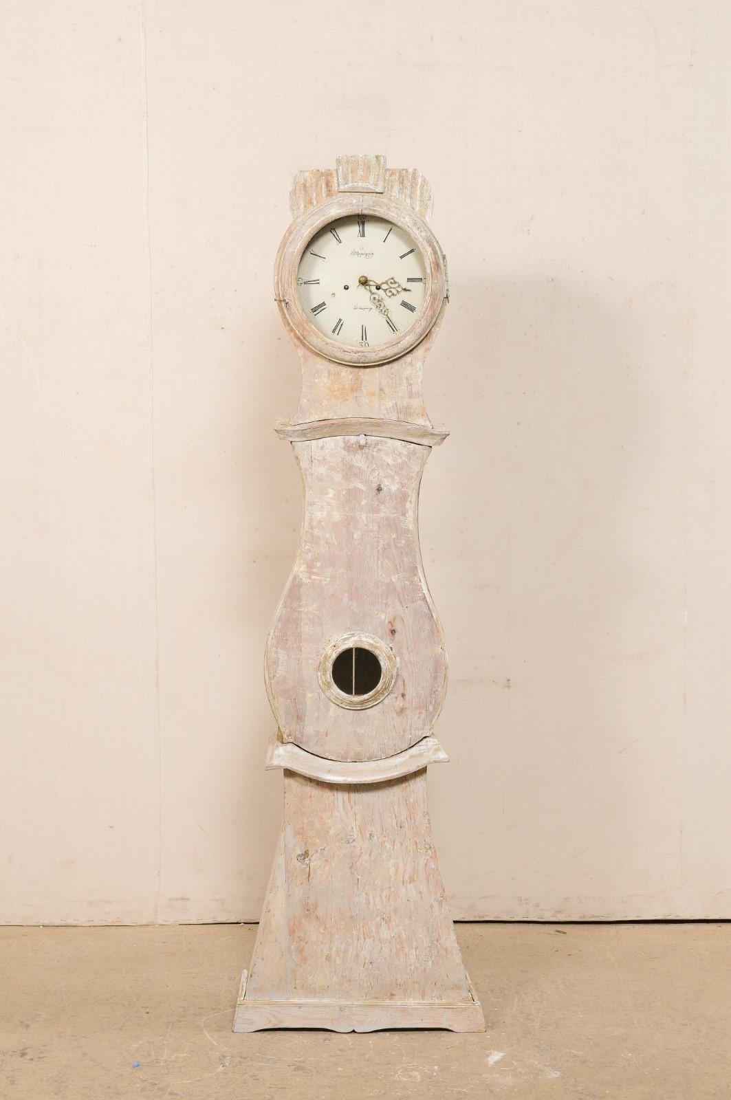 An early 19th century Swedish grandfather clock with scraped finish. This antique floor clock from central Sweden is from the 1820's and features a carved & raised crest, its original round metal face & movements, a raindrop shaped belly, a curvy