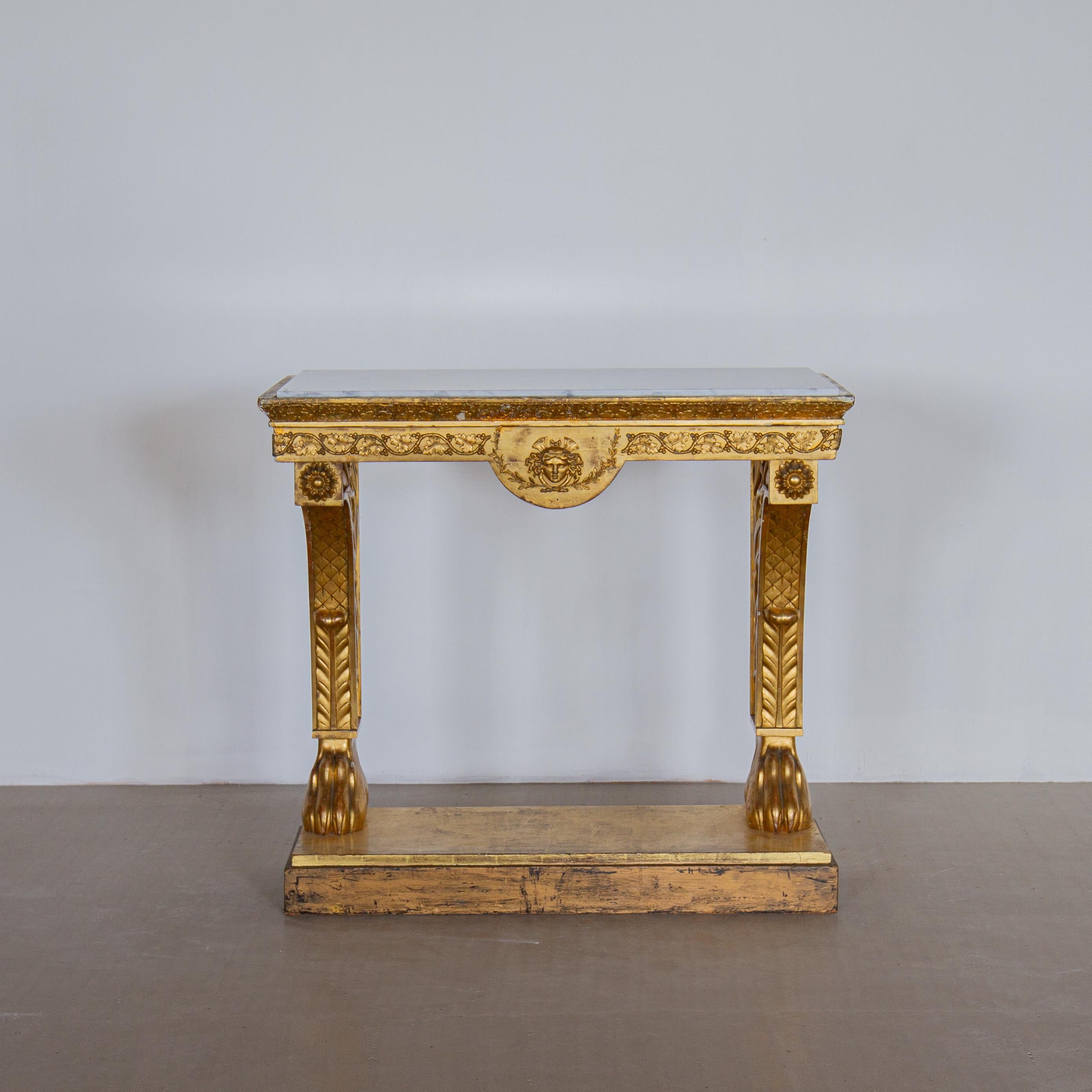An early 19th century giltwood console table with foliage and berries frieze and a central carving of medusa, on two legs set on paw feet and base and with inset veined white marble top. 

Light damage and old repairs.