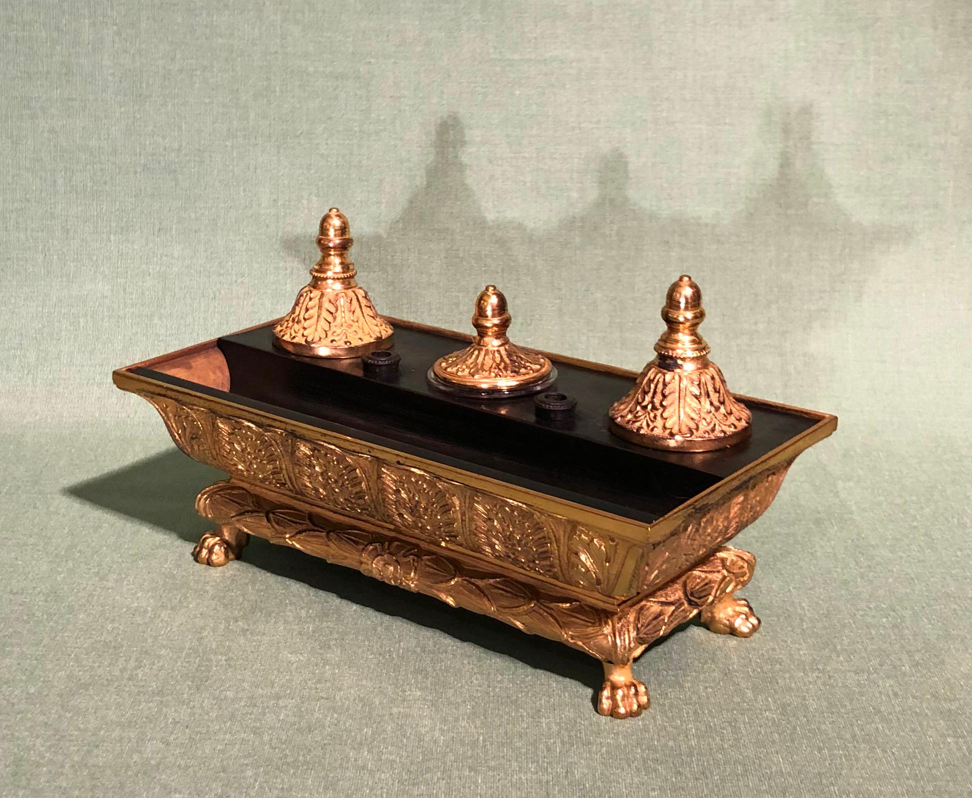 An early 19th century Regency period bronze and ormolu encrier, having penholders and triple inkwells with leaf cast lids contained in leaf decorated classical bath, supported on wreath moulded plinth ending on lion 's paw feet.