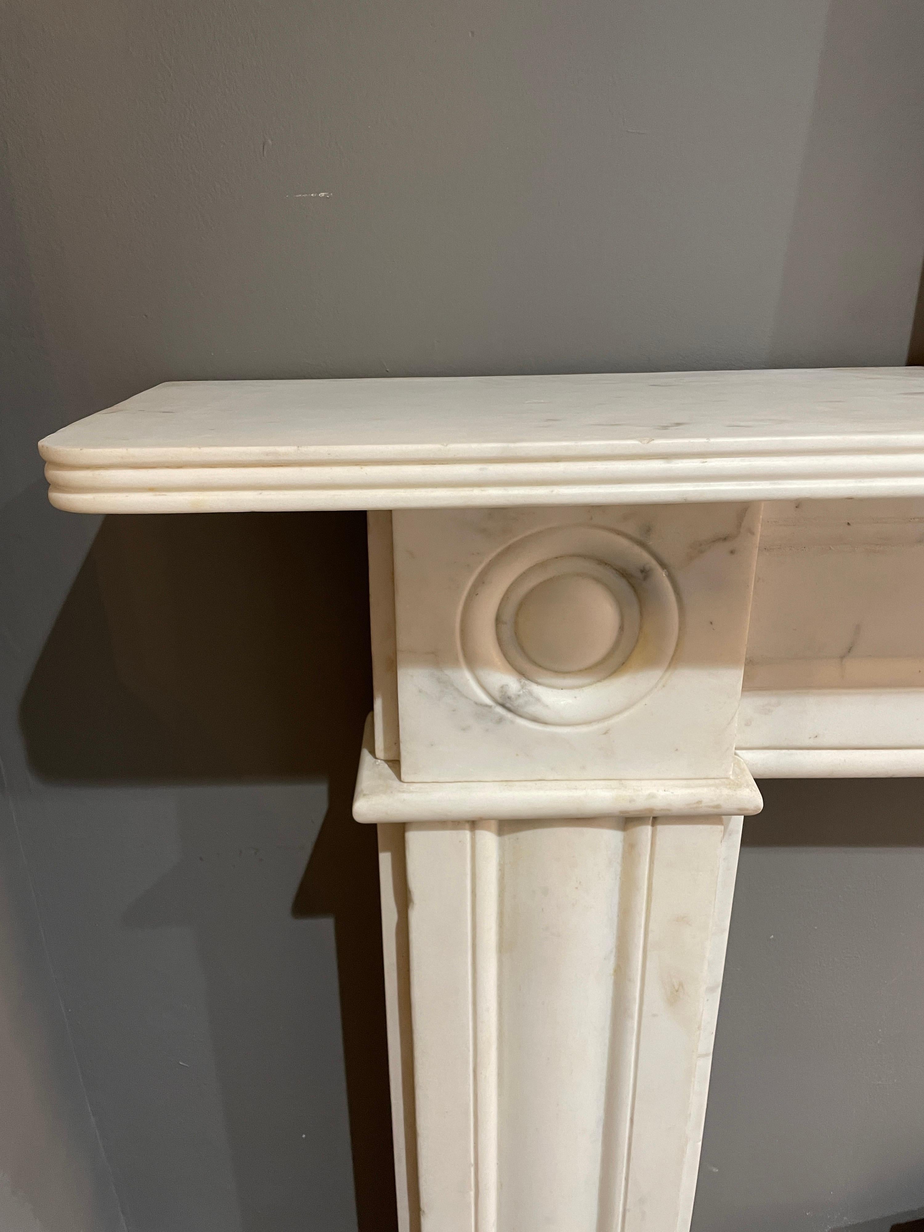 An early 19th century Regency fireplace from Dublin Ireland, executed in Italian Statuary white marble. The jambs and frieze with conforming wide mouldings, larger than usual carved roundel corner blocks and stood on separate foot blocks. All