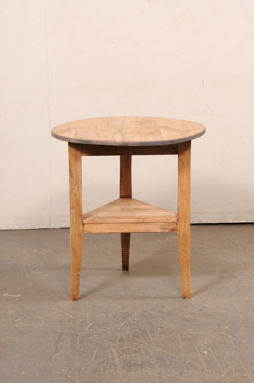 An English bleached wood cricket table with lower shelf from the early 20th century. This antique table from England features the typical cricket design with round-shaped top, which rests above an inset and triangular shaped apron, and is raised on