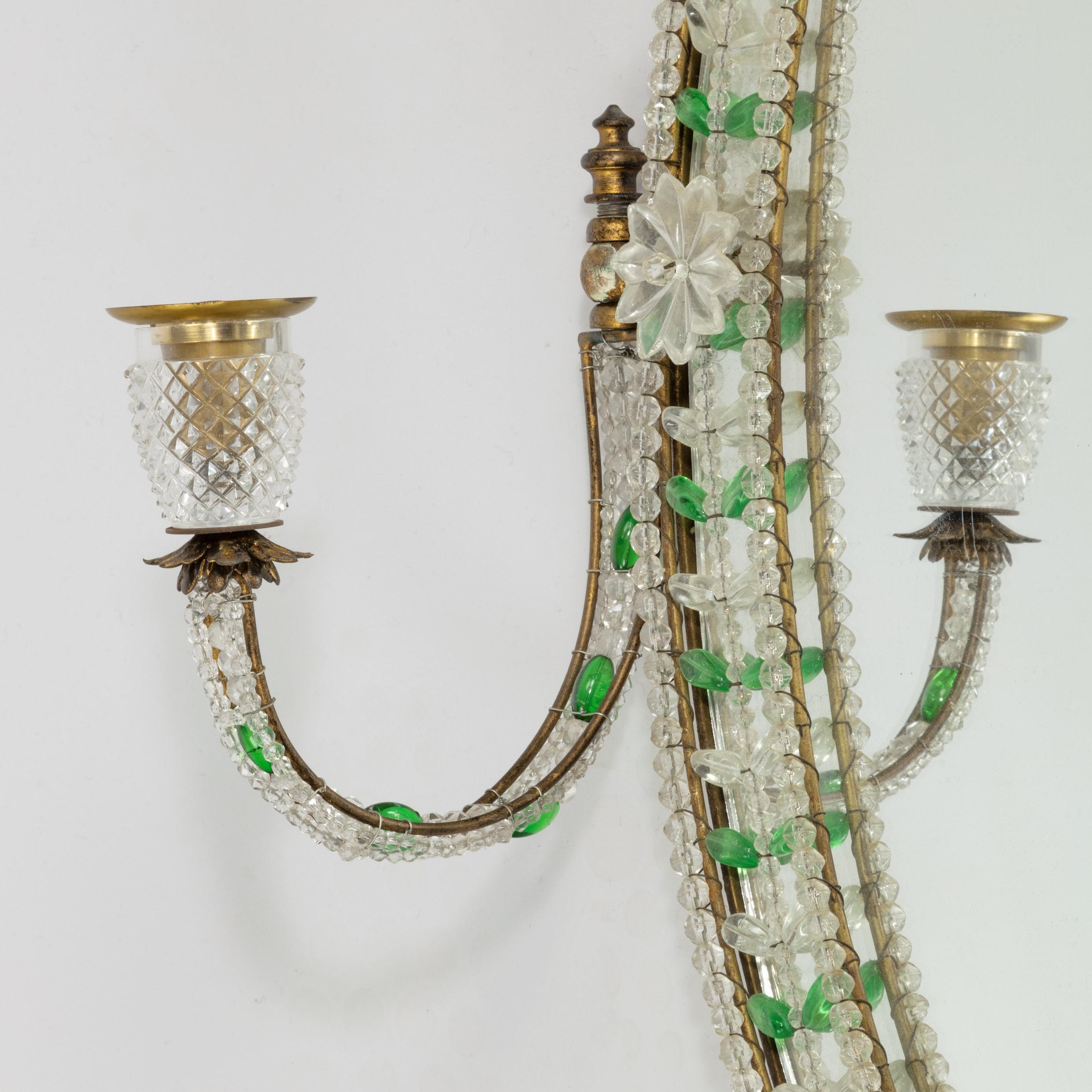 An early 20th century glass mirror, the oval plate within an ormolu frame bearing two adjustable candle arms, decorated with wire-strung clear and green glass beads and applied cut glass flower heads and sconces, English, circa 1920.
 