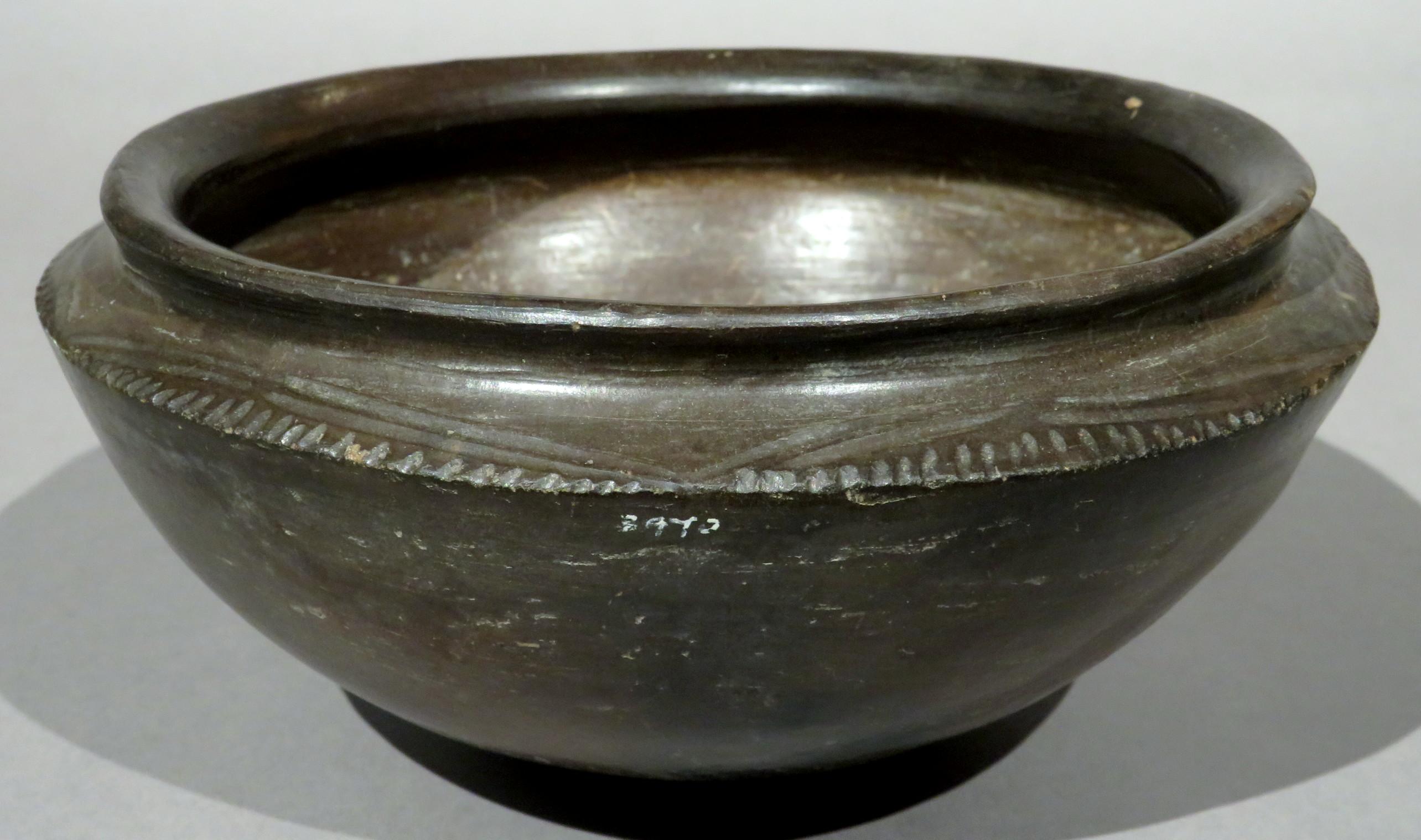The dark brown earthenware bowl-shaped body rising to a flattened shoulder with incised linear & notched detail rising to a roll-over rim. The underside showing evidence of exposure to open flame. Bearing hand painted collection inventory numbers