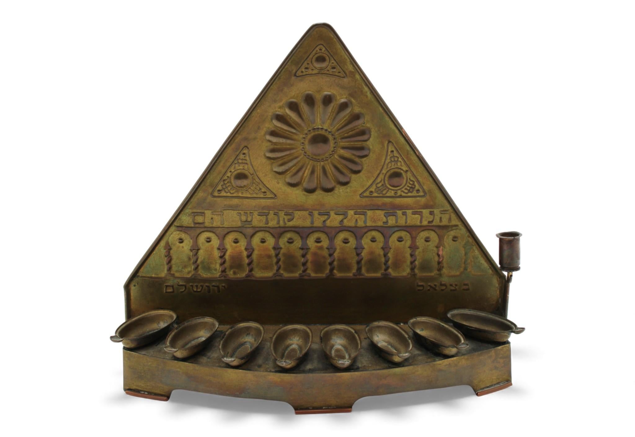 Una classica lampada Hanukkah in ottone della Scuola Bezalel, finemente lavorata con i caratteristici repoussé, elementi applicati e motivi architettonici. La panca anteriore sostiene otto coppe dell'olio ovali e allungate, fissate ordinatamente in