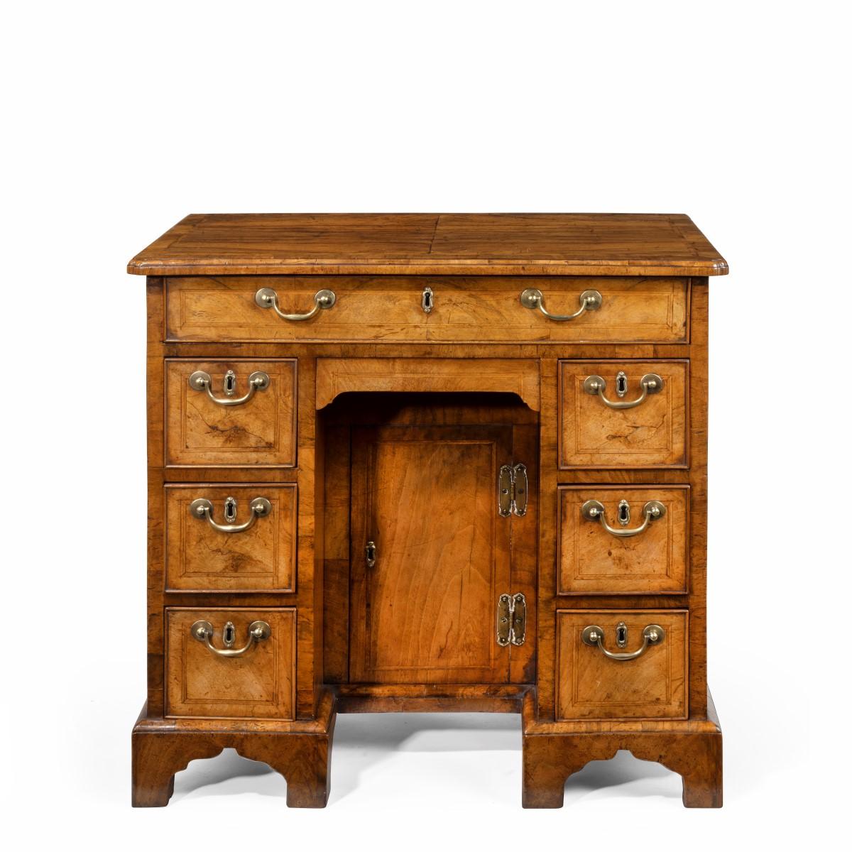 An early George III walnut kneehole desk, the shaped rectangular top quarter veneered with re-entrant corners and herring bone cross-banding, with one long drawer above a disguised drawer in the apron, the kneehole with a central cupboard and