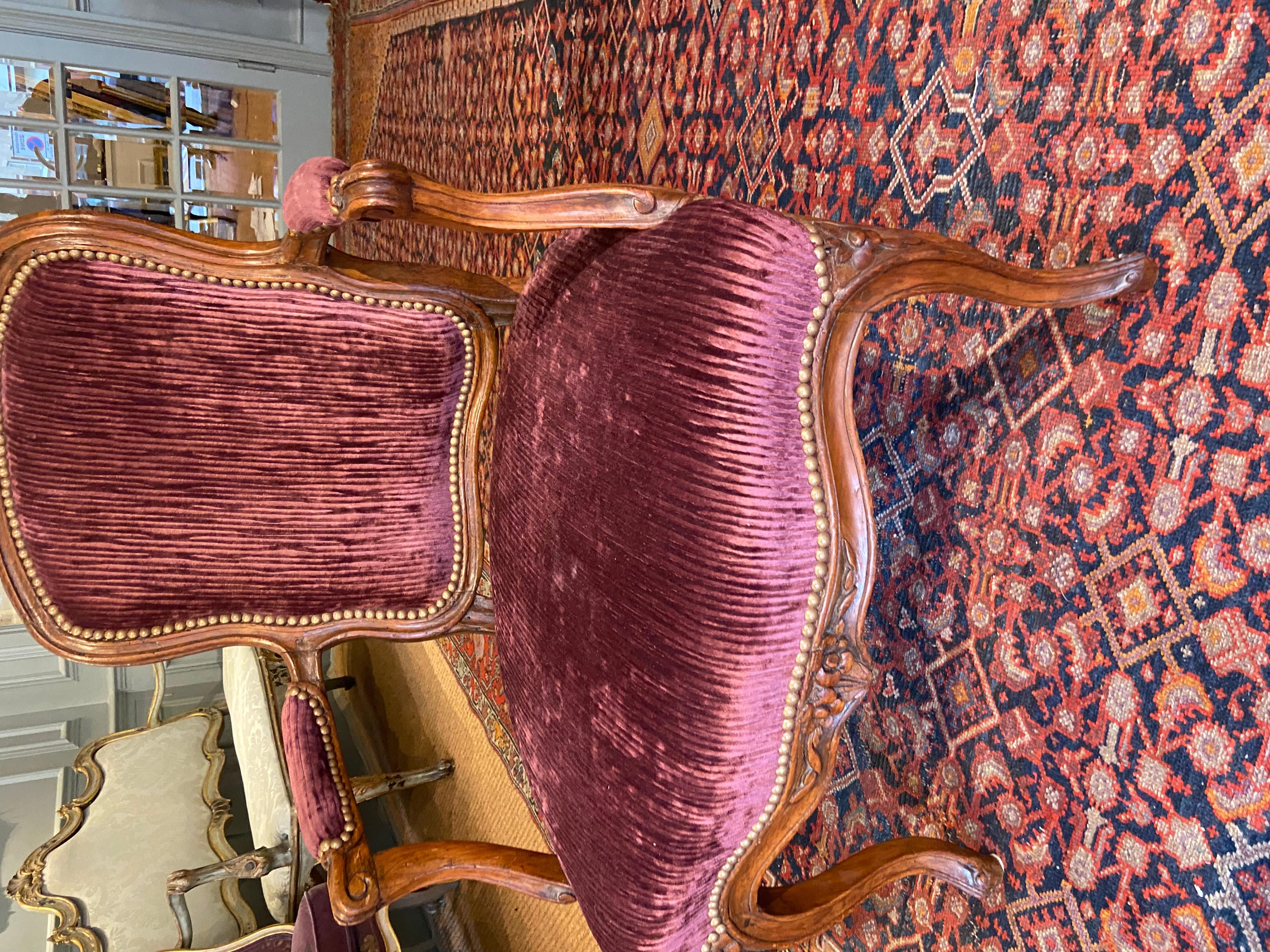 An early Louis XV beechwood fauteuil (open armchair) (mid-18th century). The cartouche-shaped padded back, arms and serpentine-fronted seat, upholstered à chassis in plum velvet, Channelled frame with central floral motif on hipped cabriole legs.