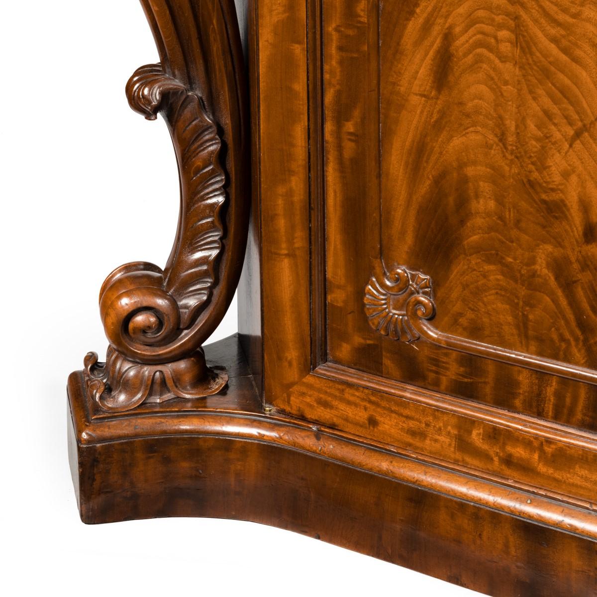 An early Victorian two-door mahogany side cabinet, attributed to Gillow, the shaped rectangular top with cut corners above two cupboard doors flanked by two deeply carved bold acanthus and C-scroll corbels, enclosing a fitted shelf, all raised on a
