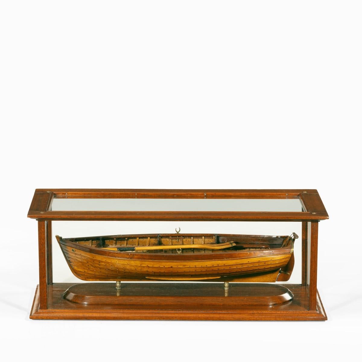 An Edwardian clinker-built model of a gentleman’s yacht tender, with a boat hook and oars, teak gratings, mahogany seats and thwarts, the rudder with a yoke, brass rowlocks and rubbing strake to the stern, within a glass case. English, circa