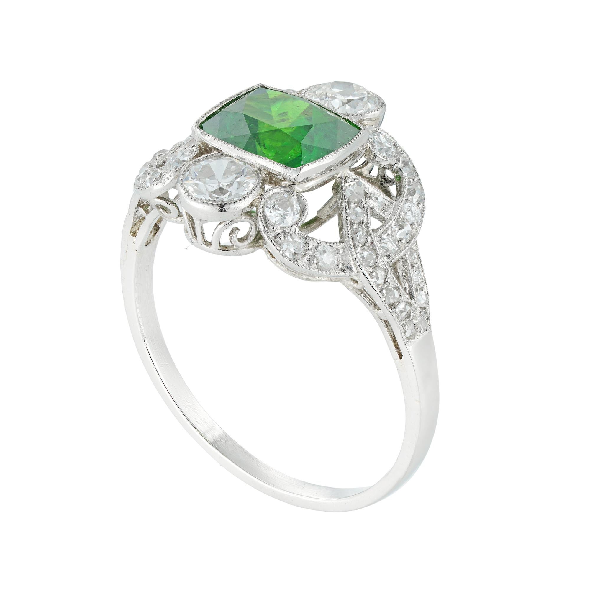 An Edwardian Demantoid garnet and diamond ring, the cushion-cut green garnet weighing 2.05 carats, accompanied by GCS report 81281-75 stating to be natural Demantoid garnet, with horsetail inclusions, vertically-set between two old European-cut