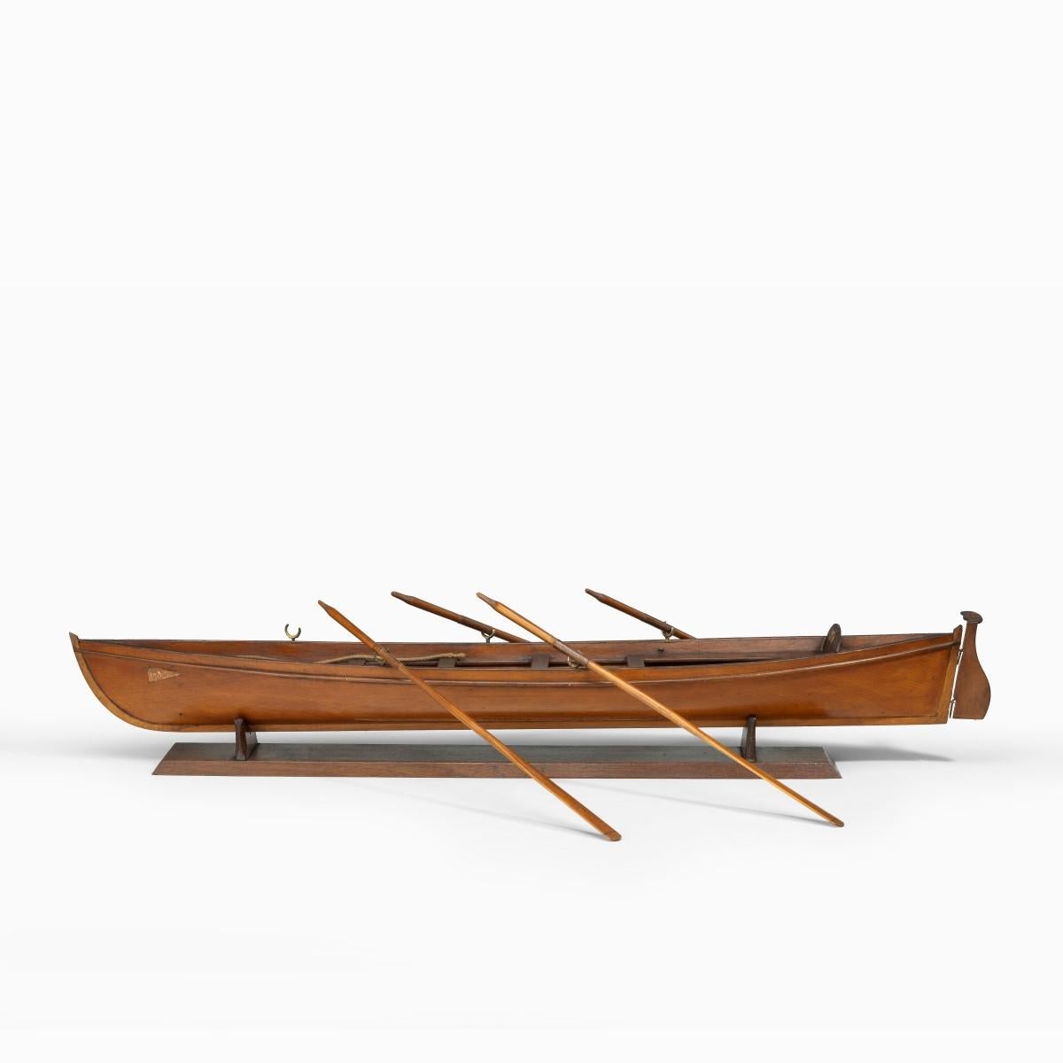 An Edwardian pine five-oar rowing skiff, retaining just four oars, but with the original mast and boat hook, a paper pennant on the back rest showing three heraldic lions, with a burgee on the bow, raised on a stand. English, circa 1900.