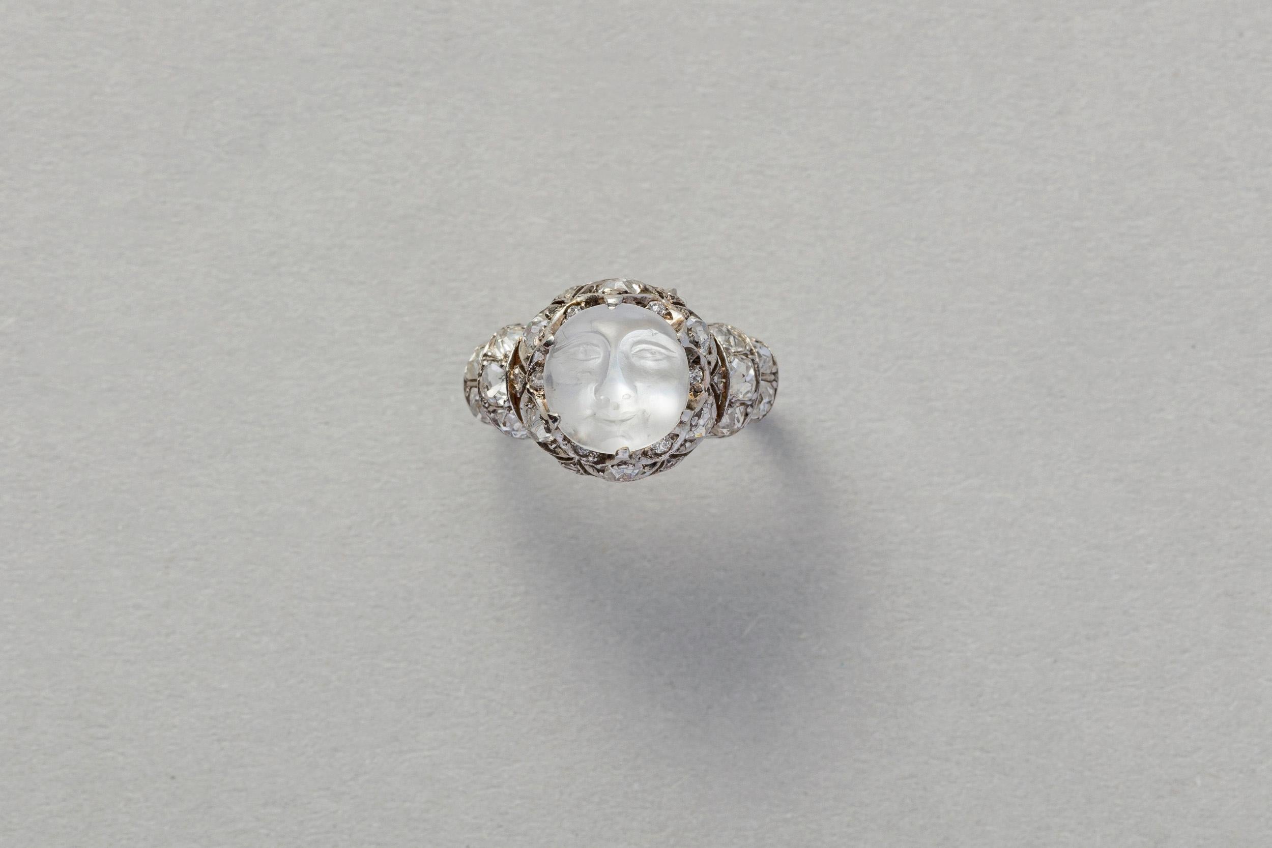 A sweet Edwardian platinum ring set with a round, silky white moonstone engraved with the face of the moon. Around and underneath the six prongs are different size and shape old cut and rose cut diamonds. On the shank of the ring are two curves one