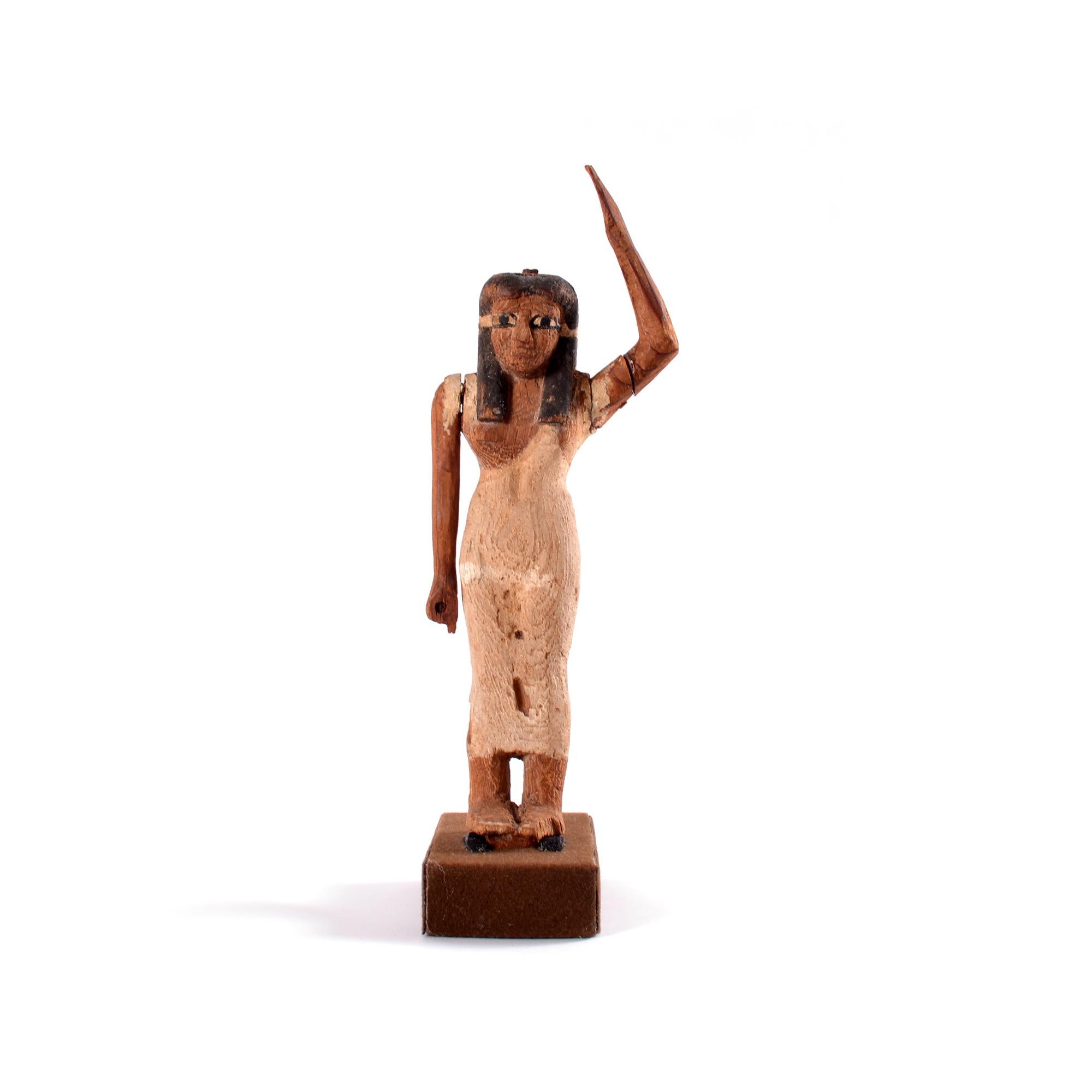 Standing female wearing a tight fitting white dress and black tripartite wig, serene facial features with large painted eyes. Separately made arms attached with dowel to the shoulder, left arm raised and brought back to support the basket of