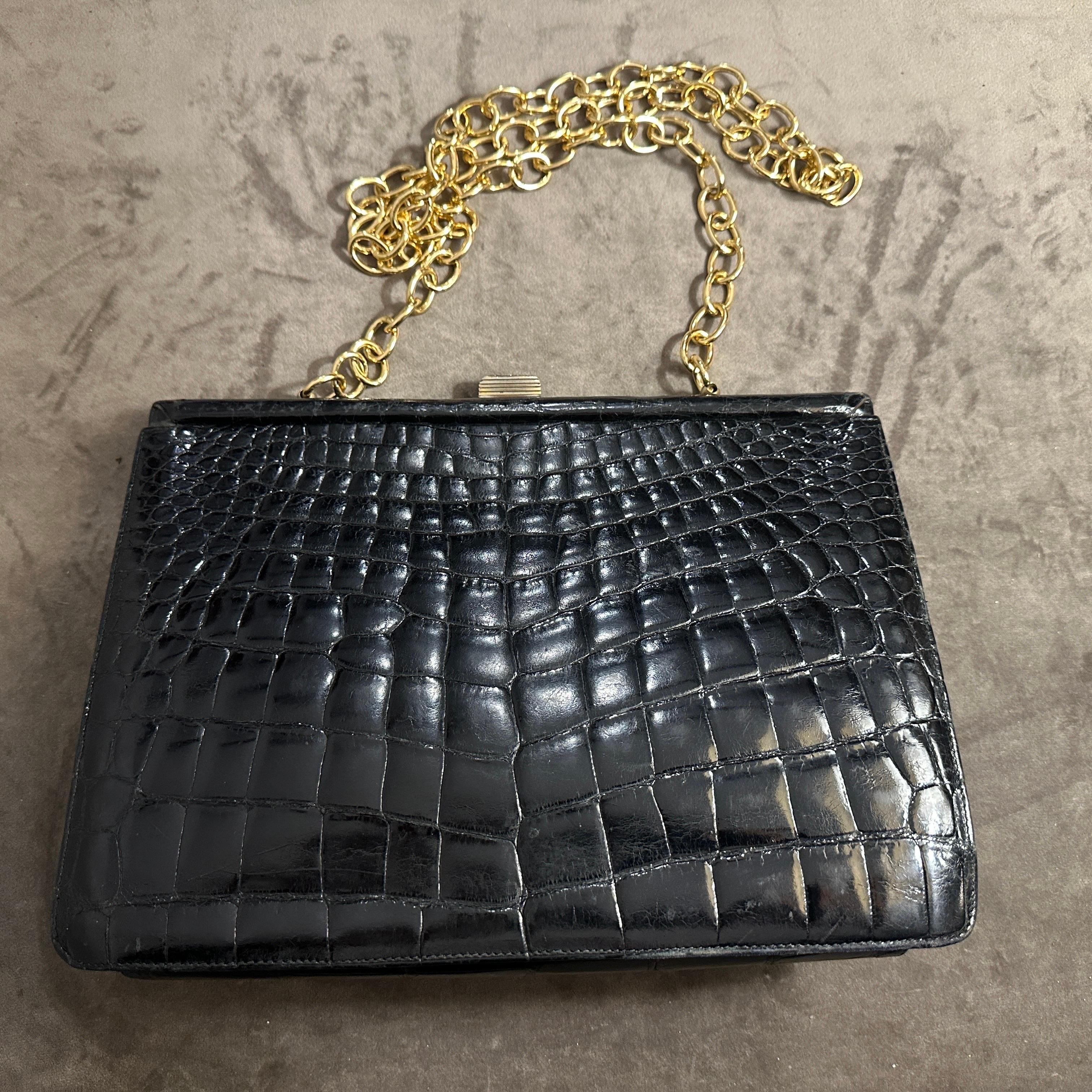 This is an elegant Italian shoulder bag from the 1950s, showcasing a luxurious combination of black leather and brass accents. The bag's exterior is crafted from genuine crocodile leather, featuring its signature glossy finish and distinctive scale