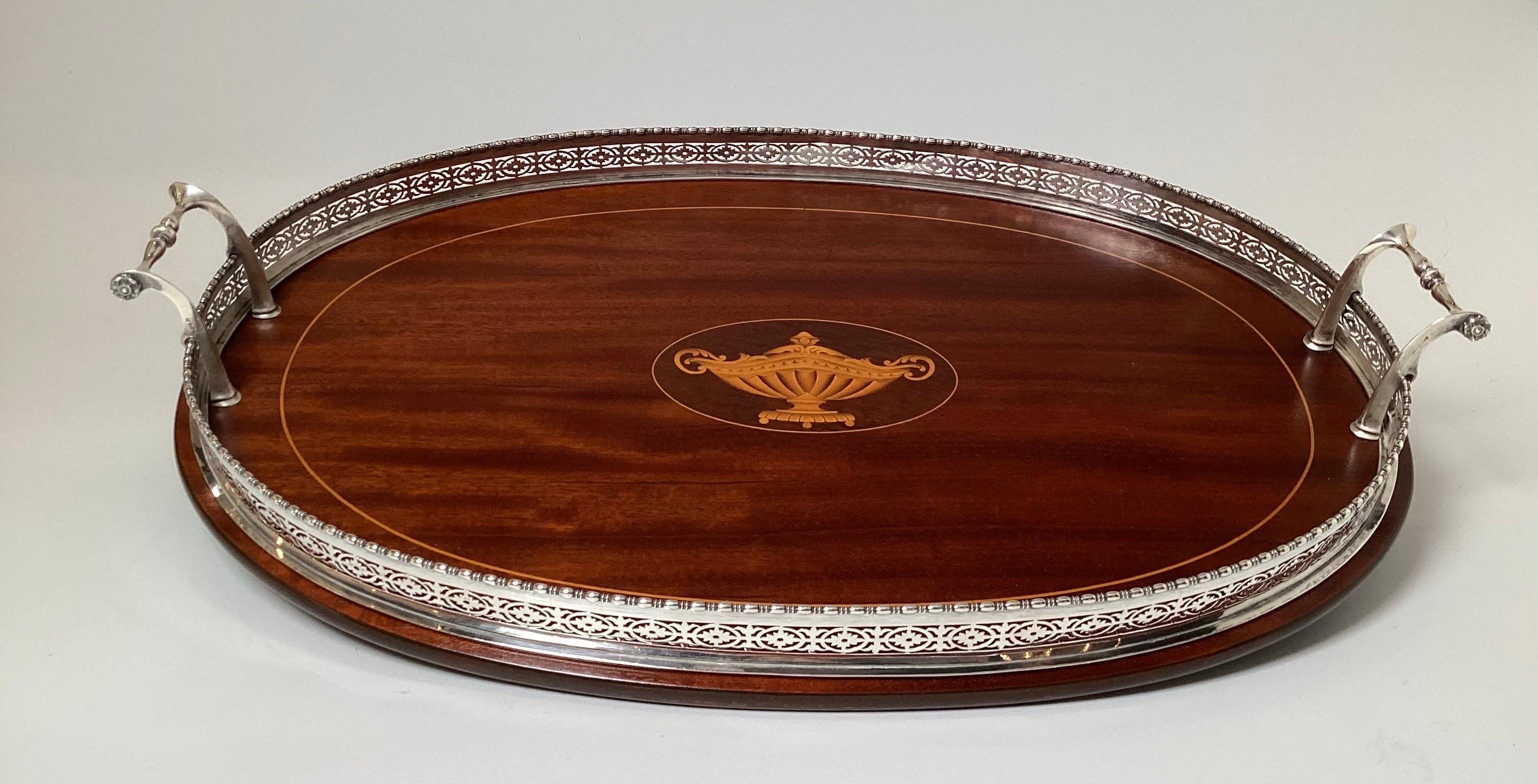 Elegante bandeja ovalada con incrustaciones de madera de caoba y satén, con asas de plata de ley y borde de galería.  Bandeja con incrustación central de motivo de urna con borde de galería de esterlina reticulada.  21 pulgadas de mango a mango, 14
