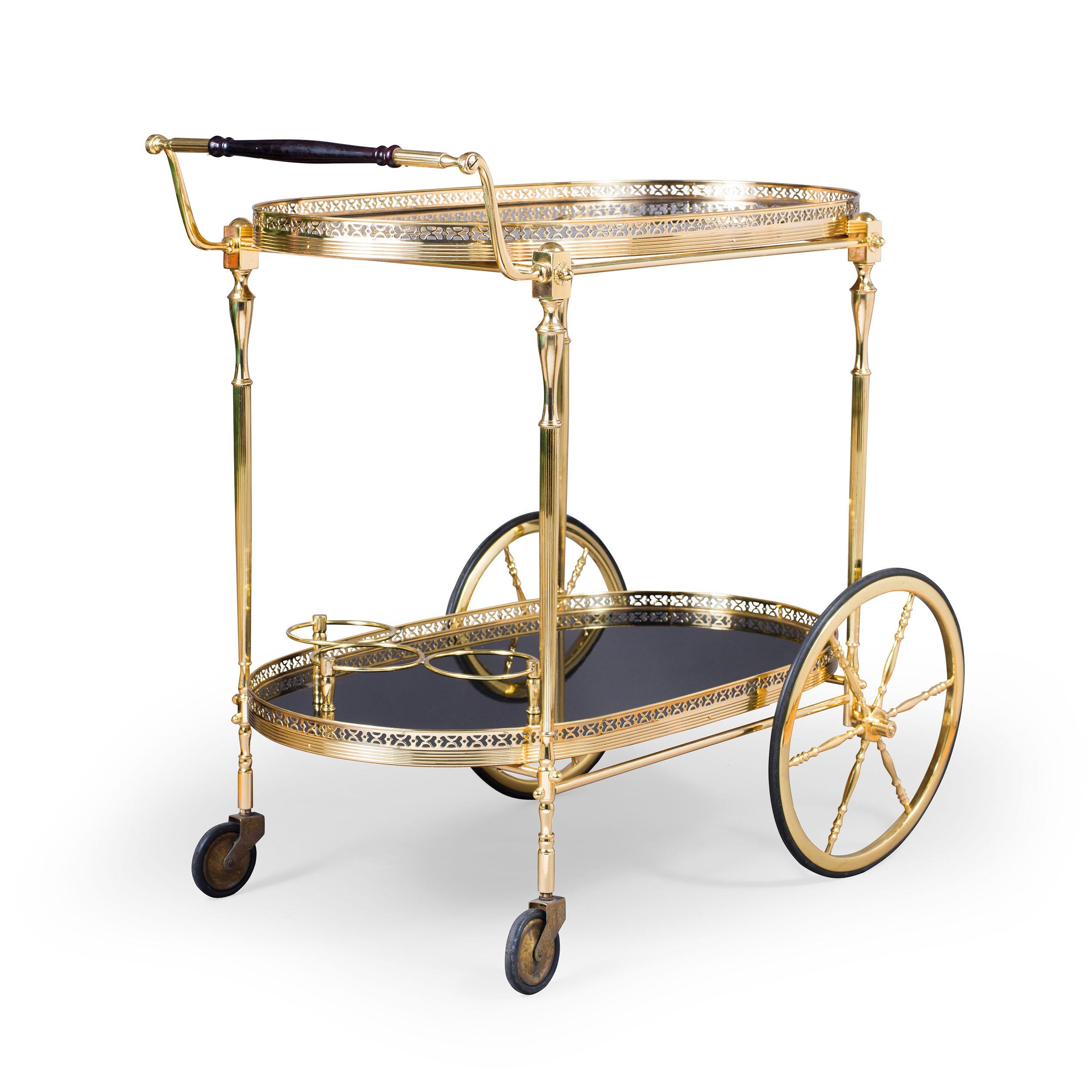 An elegant mid-century drinks trolley with two oval tiers, both with lacquered black surfaces, framed with pierced brass galleries, and supported on turned brass legs with cast brass decorative details applied. Large turned brass wheels at the