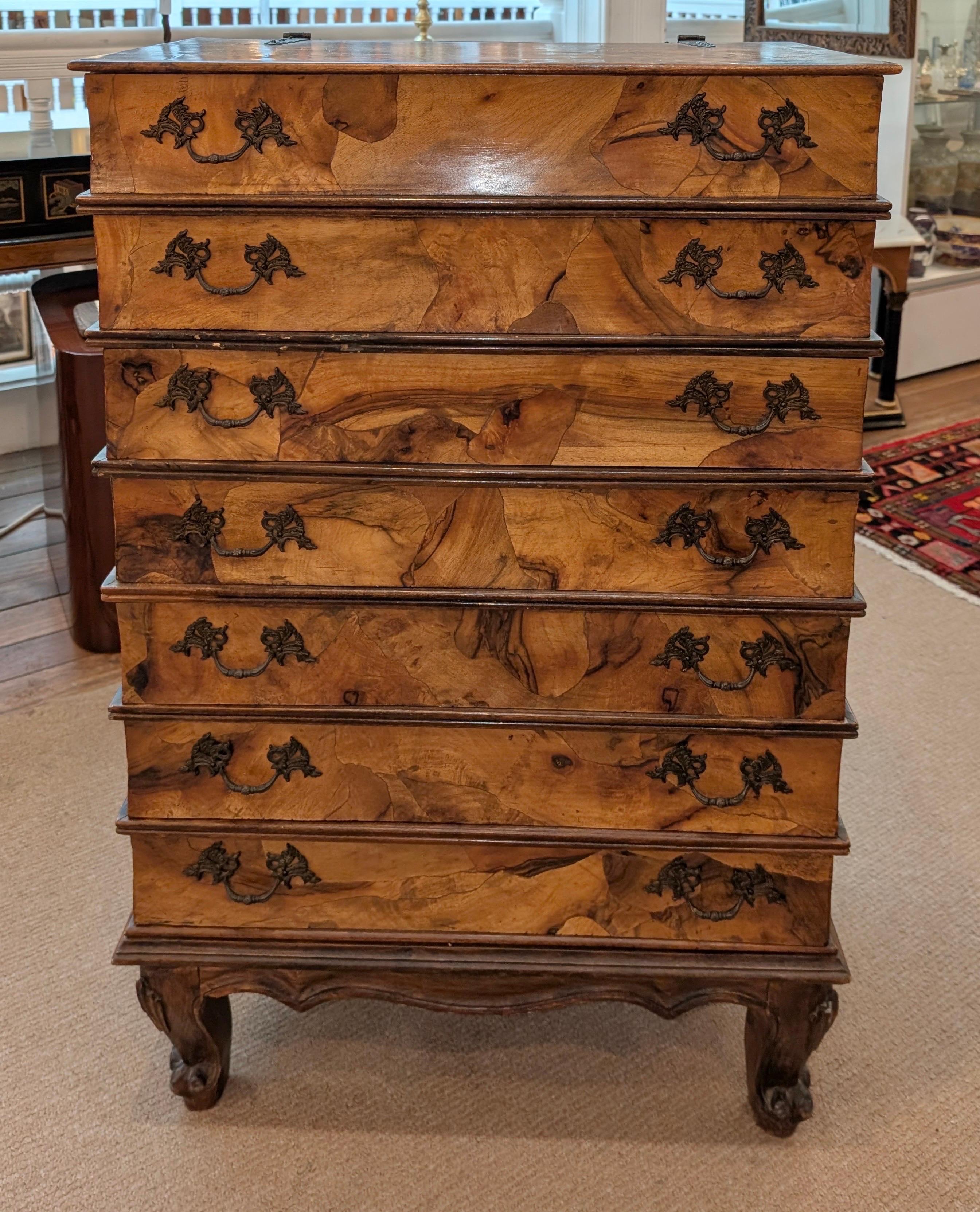 Una cassettiera italiana in legno d'ulivo, circa 1950.  La cassapanca ha la forma di una pila di libri per un totale di 7, di cui 6 cassetti e il piano con un pannello apribile.  Il legno altamente figurativo in toni medi poggia su quattro gambe