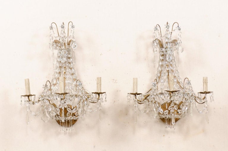A French pair of three-light crystal wall sconces from the mid-20th century. This pair of vintage wall candelabras from France each feature an upper waterfall crown adorned with glass-cut flowers and crystals, with surmounting cascades of delicately