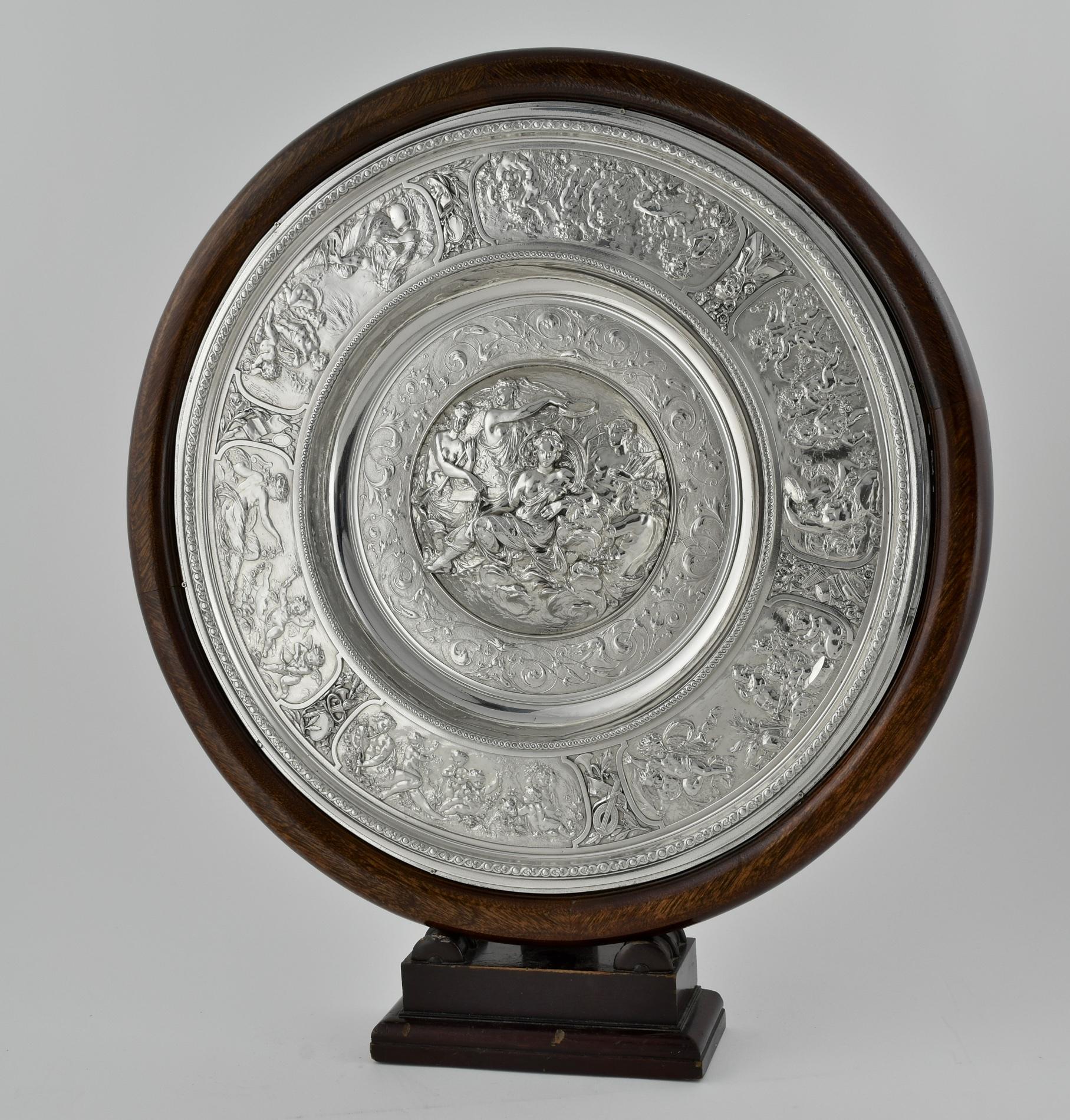 A shield by Elkington & Co decorated with classical scenes. The centre is embellished with classical female figures playing musical instruments. Continuing with the musical motif, around the edge  of the shield is decorated with scenes of musicians