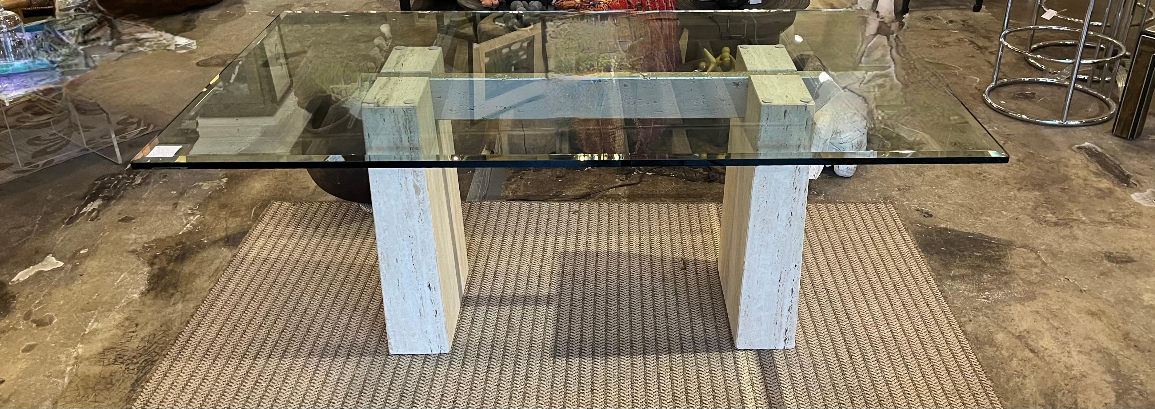A 1972 travertine and beveled glass dining table designed by Guy Barker for Ello Furniture. The sustantial travertine marble pedestals uphold a glass top with almost one inch surrounding the perimeter. The custom under beam has been painted a