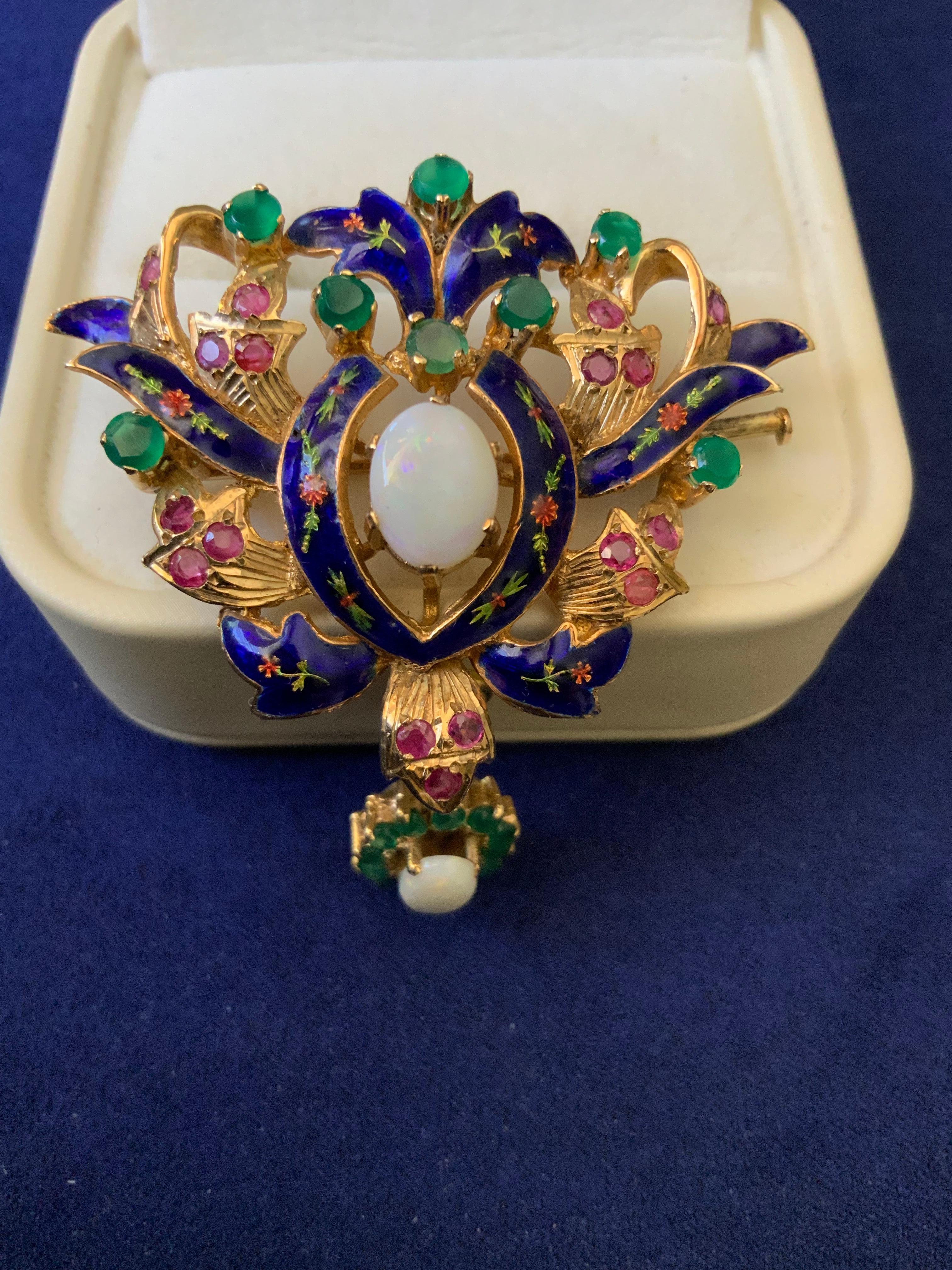 This is an Emerald Opal Ruby Blue Enamel Gold Brooch. It depicts a shield like gold brooch with gold branches forming a wreath decorated with a blue enamel flowers bows. An oval Opal stone enhances the center of the wreath and rubies and emeralds in