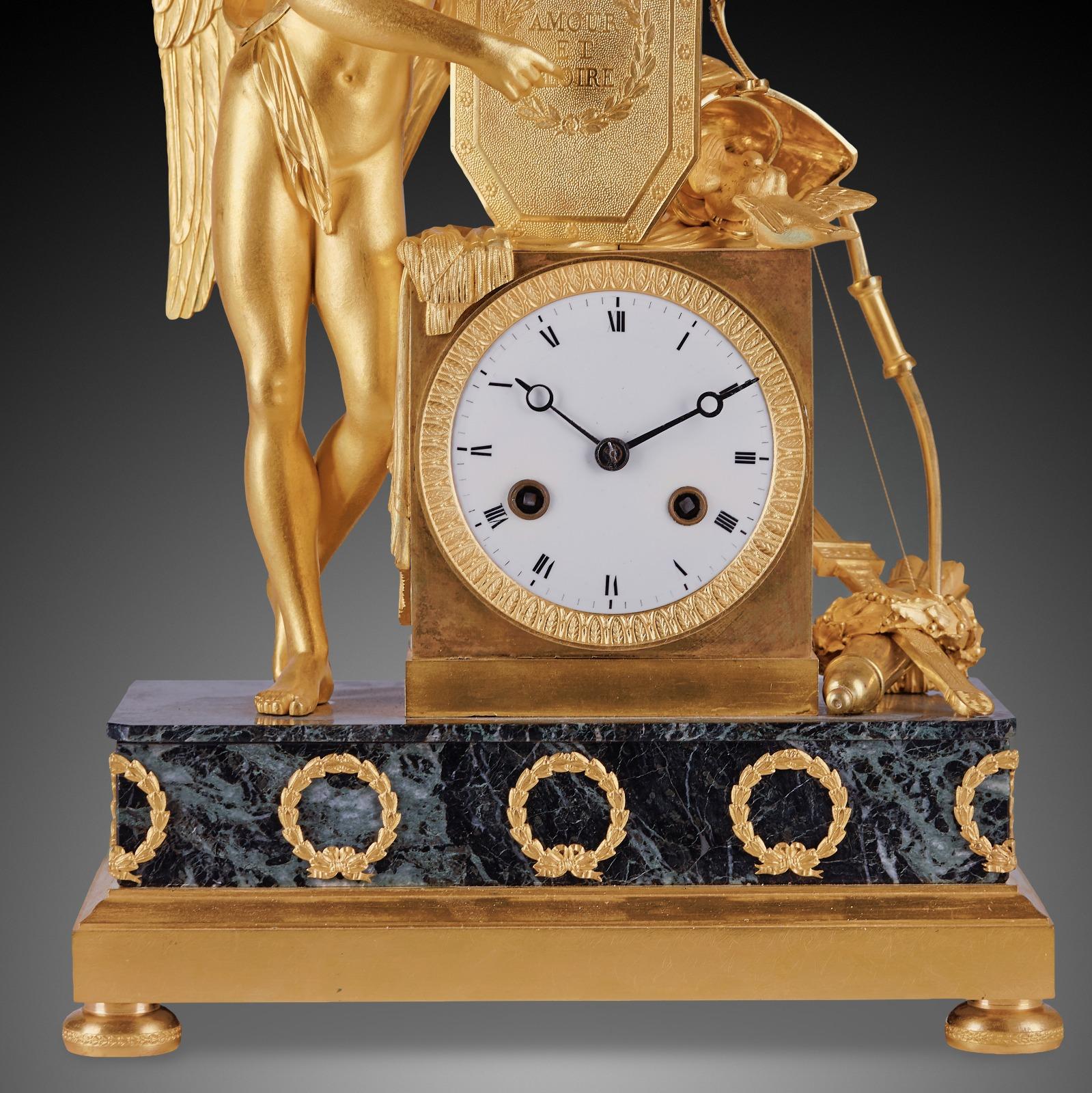 An Empire Ormolu And Verde Antico Mantel Clock
The circular white-enamelled Roman dial sigend within a rectangular case surmounted by a winged helmet and cooing doves, flanked by a winged youth holding a shield inscribed 'AMOUR ET GLOIRE', on a