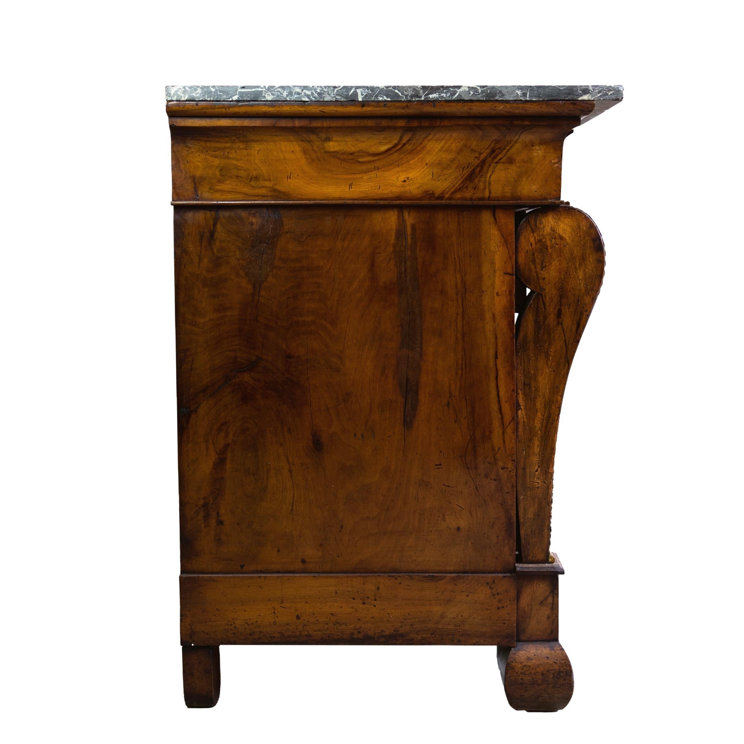 An Empire Commode in figured mahogany with a Belgian black fossilized marble top above a frieze drawer and three uniform drawers flanked by two squared columns, each with ormolu mounts in the form of stylized acanthus leaves to the knees, the pulls