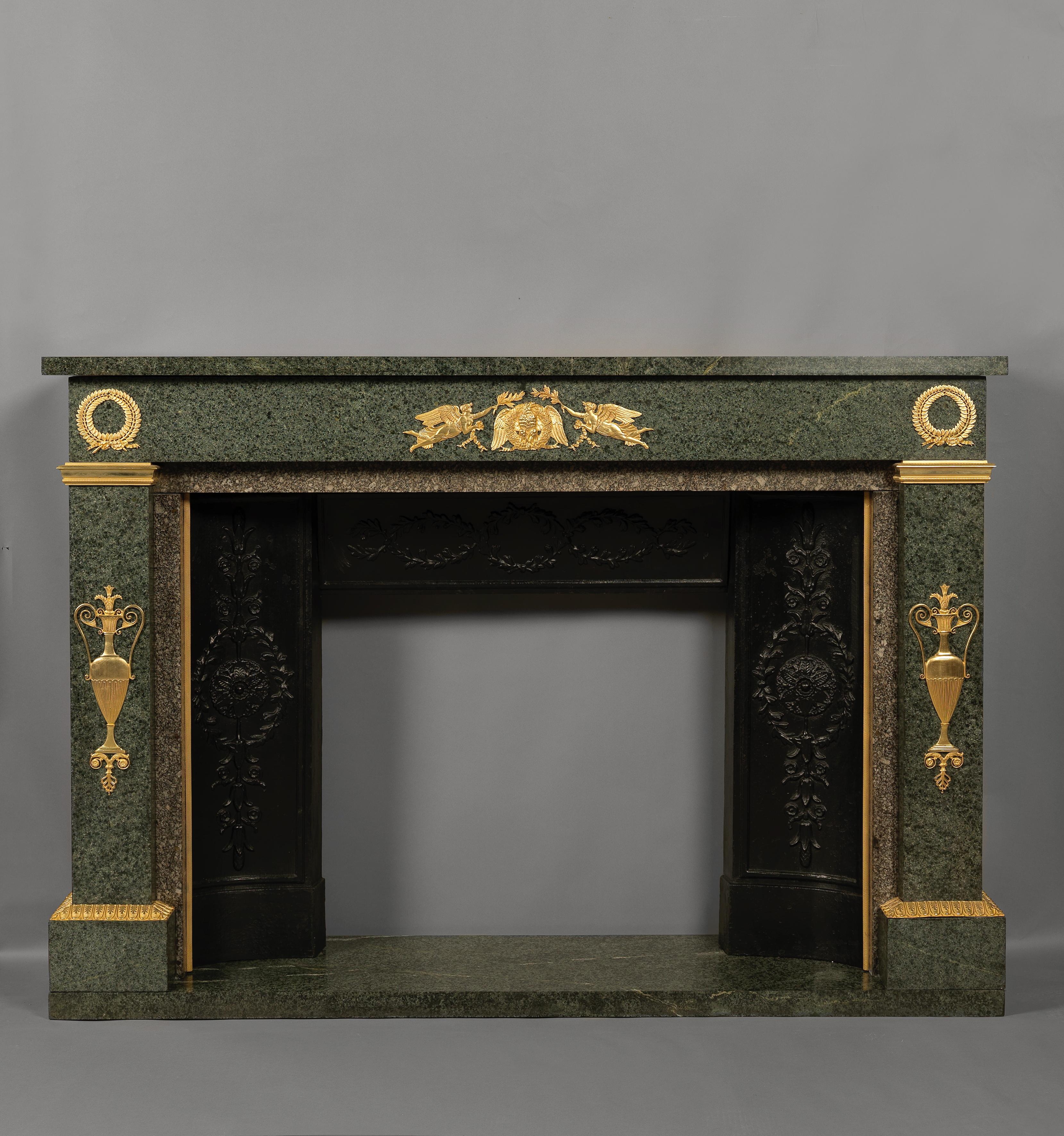 An Empire style gilt bronze-mounted green granite fireplace. 

French, circa 1850. 

The fireplace has a rectangular mantel shelf above a frieze centred by applied gilt-bronze mounts of an Eagle and wreath supported by winged female figures