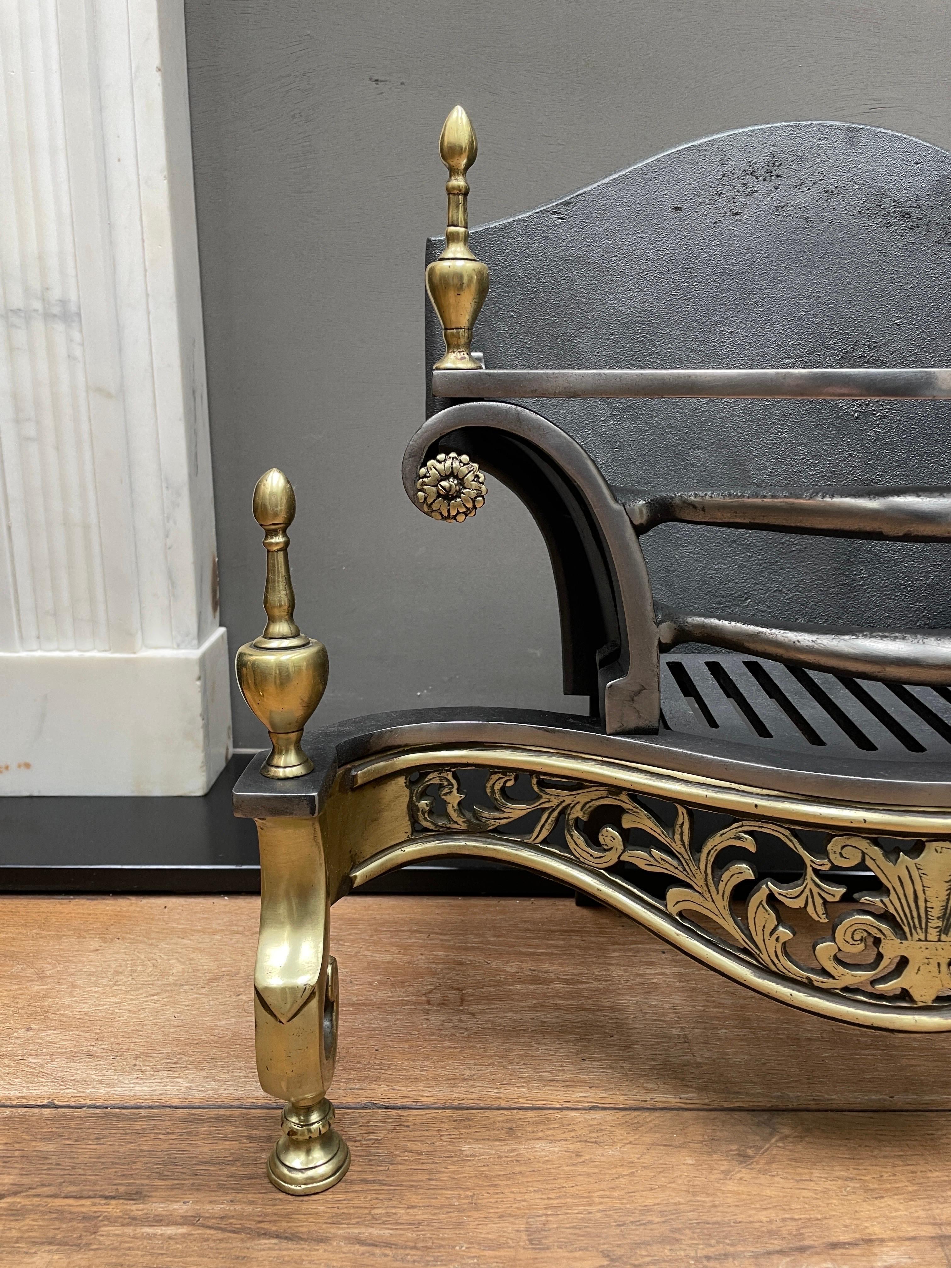 An English brass and wrought iron fire grate. The scrolled and shaped legs surmounted by finials, flanking a curved and cut brass fret of foliage and central feather. The wrought iron shaped bars again with matching finials and rosettes. 

English