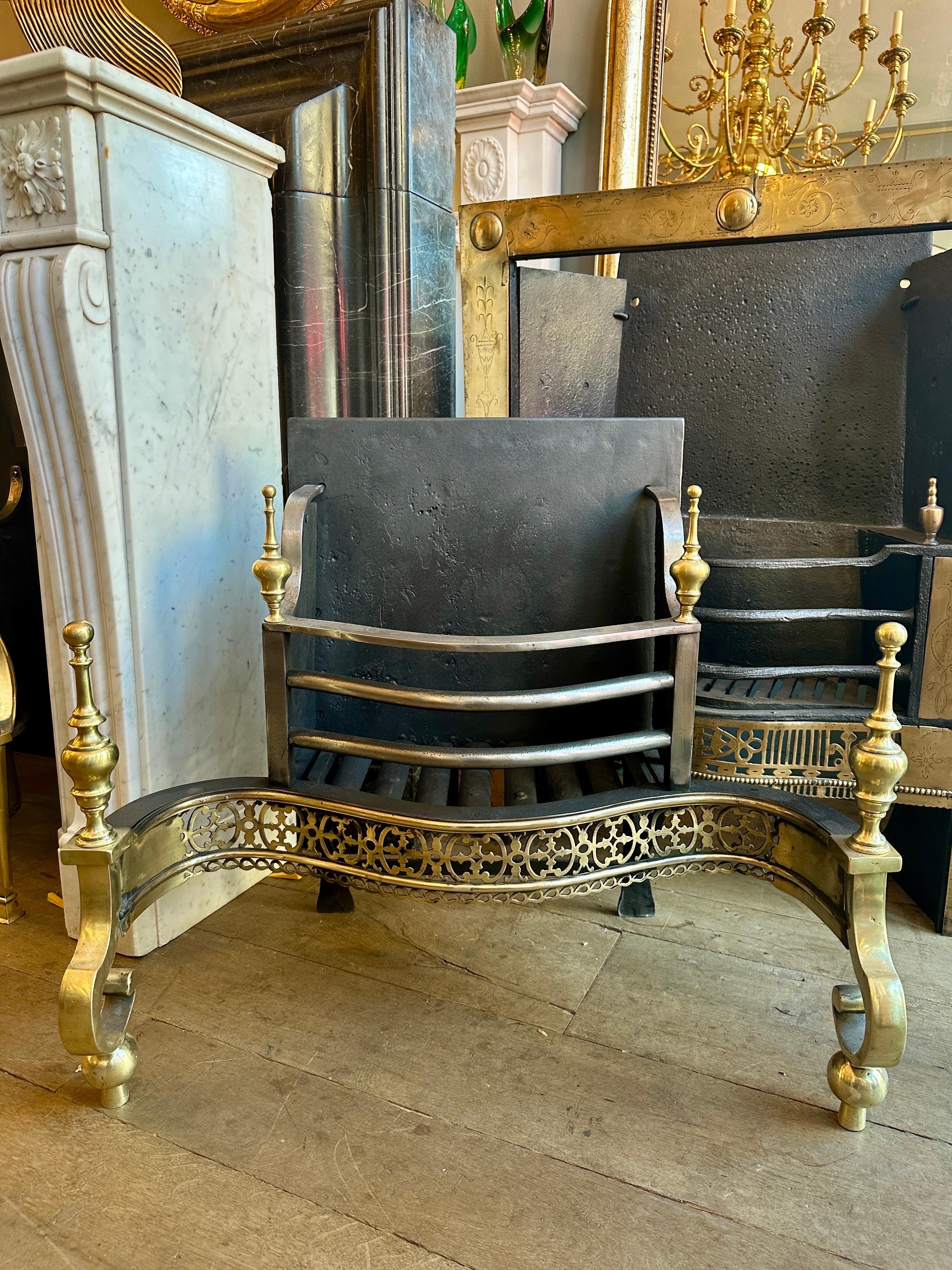 A large 19th century George II style Dog grate in steel and brass. The broken front leg supports capped with steeple finials, flanking a decorative cut brass fret with geometric apron. The bowed stele burning are again topped with smaller steeple