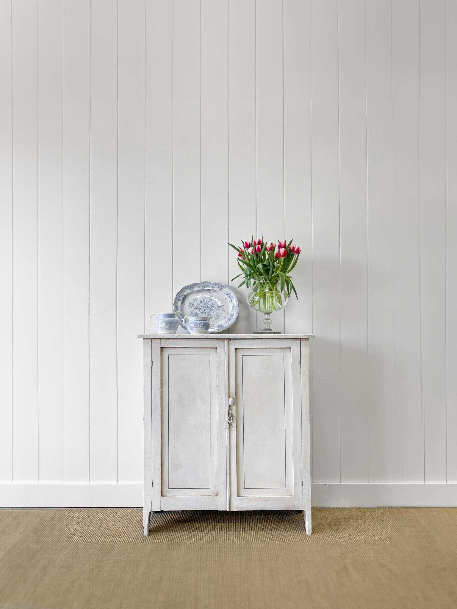A beautiful cabinet with worn white paint. A delicate line of blue provides a lovely accent. Wear in all the right places. Intricate key hold cover and beautiful hardware. Simple solid stick feet and paneled doors. Useful shelving inside.