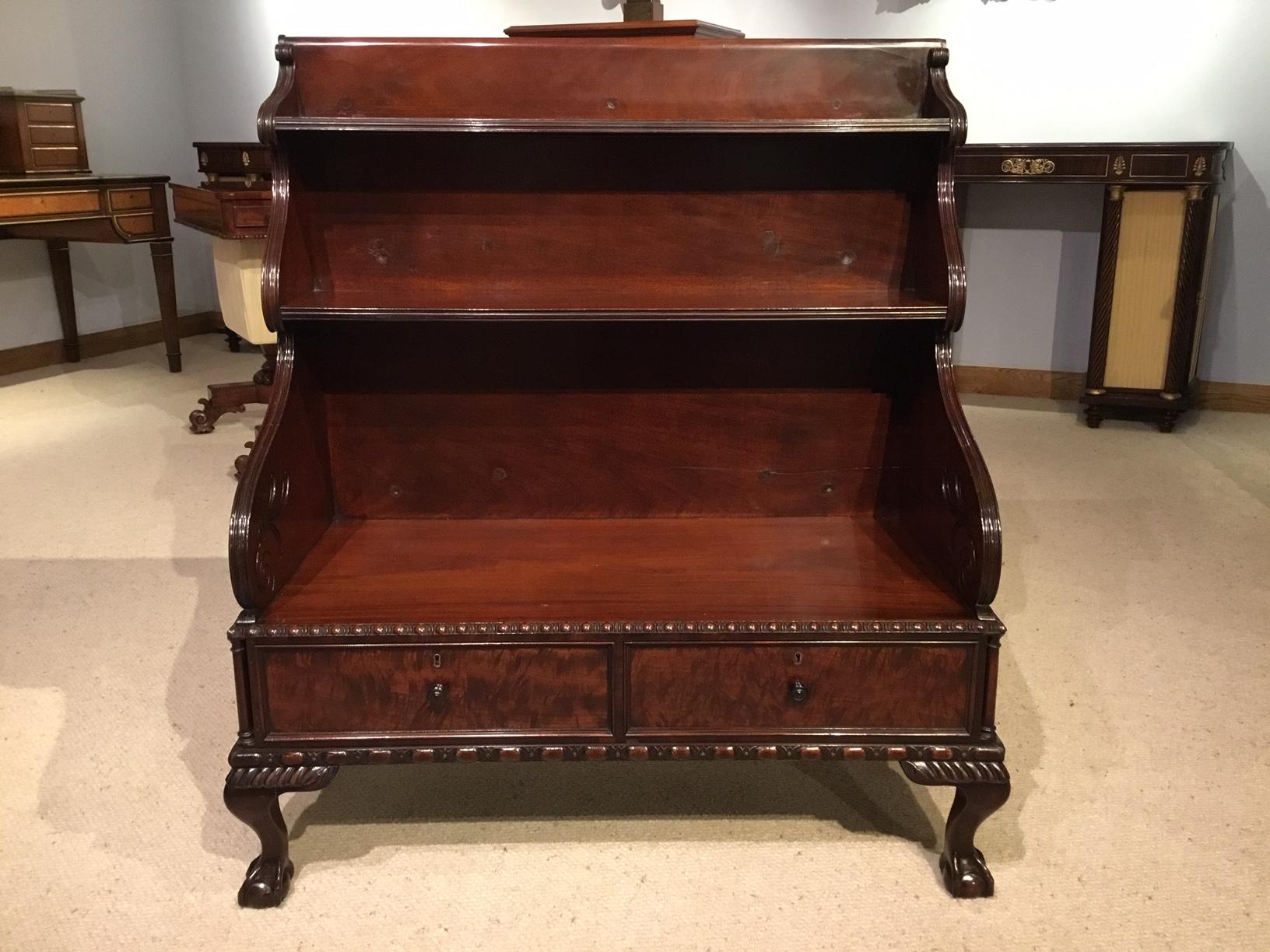An English mahogany Regency Period waterfall bookcase. Having a Cuban mahogany back panel and two raised shelves with reeded edges, finely carved shaped ends and two mahogany line drawers to the base. The drawers veneered in the finest figured