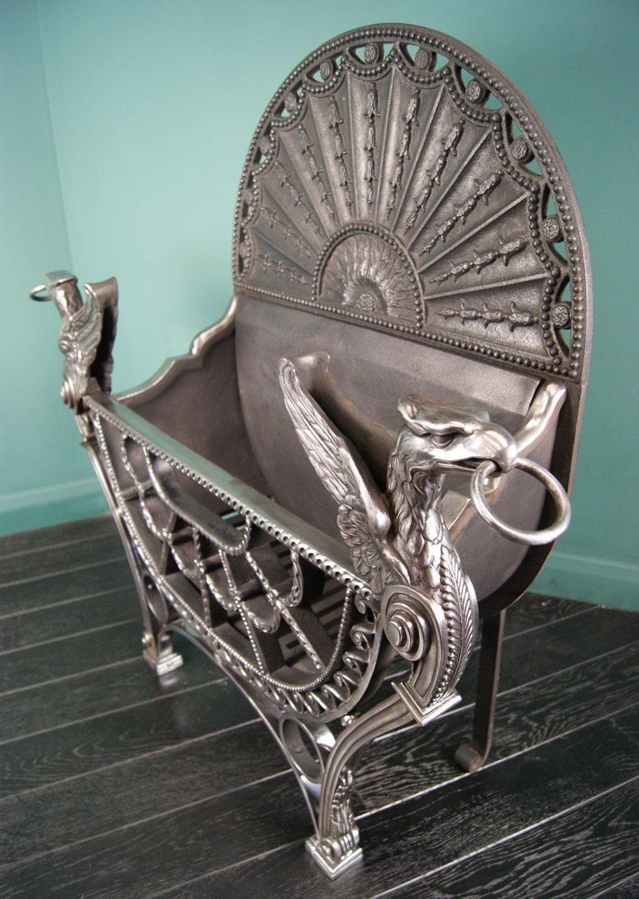 A large English polished wrought and cast-iron fire grate in the manner of Robert Adam. The openwork fan-fronted basket is flanked by winged eagles, over wrought circles with plinthed feet. The beaded arched fire back with bluebell drops in between