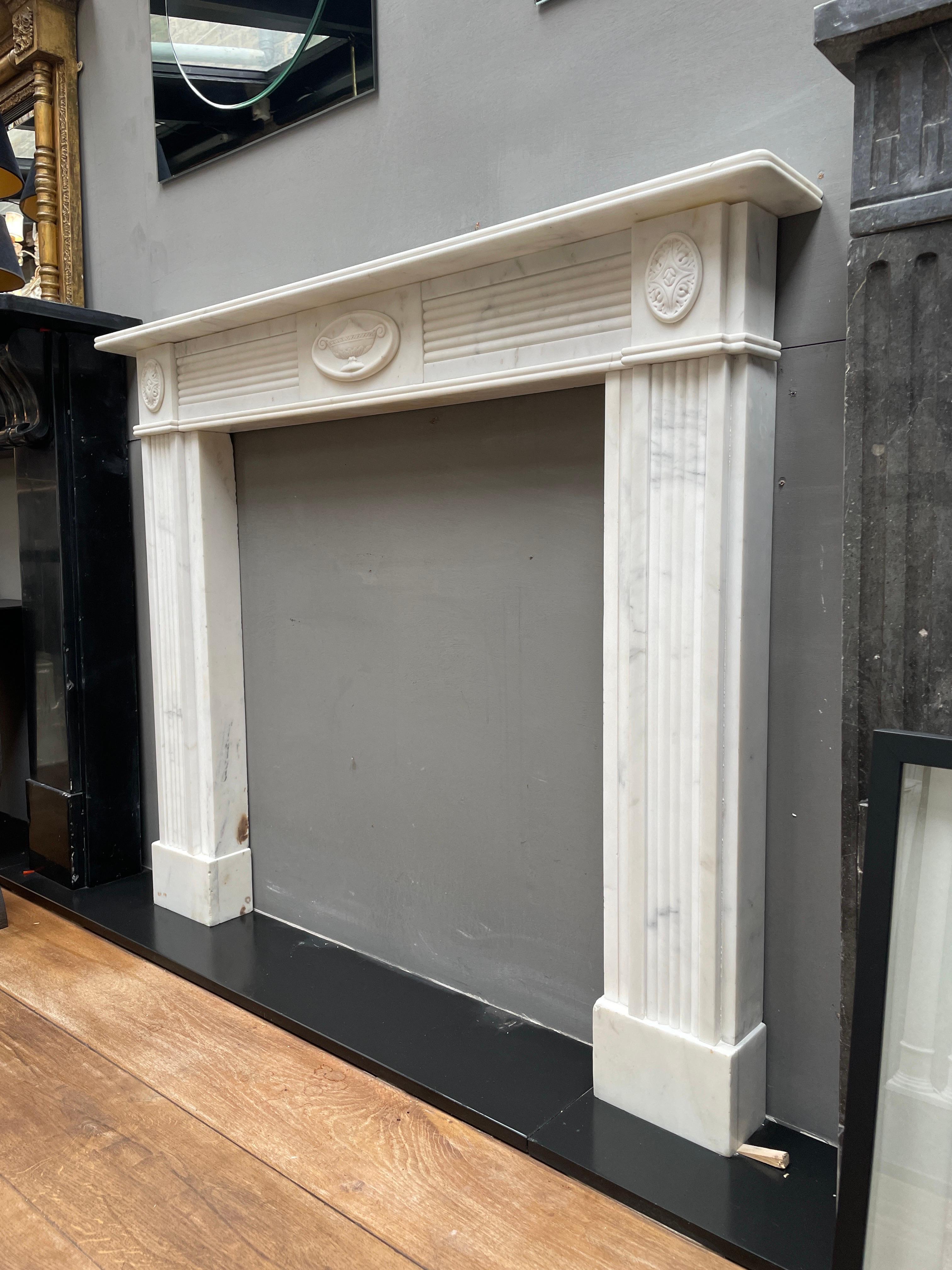A very smart and compact early 20th century Regency style fireplace in statuary white marble. The jambs with reeded front panels supported on square foot blocks, terminating in carved patarae corner blocks. The reeded frieze with centre tablet of a