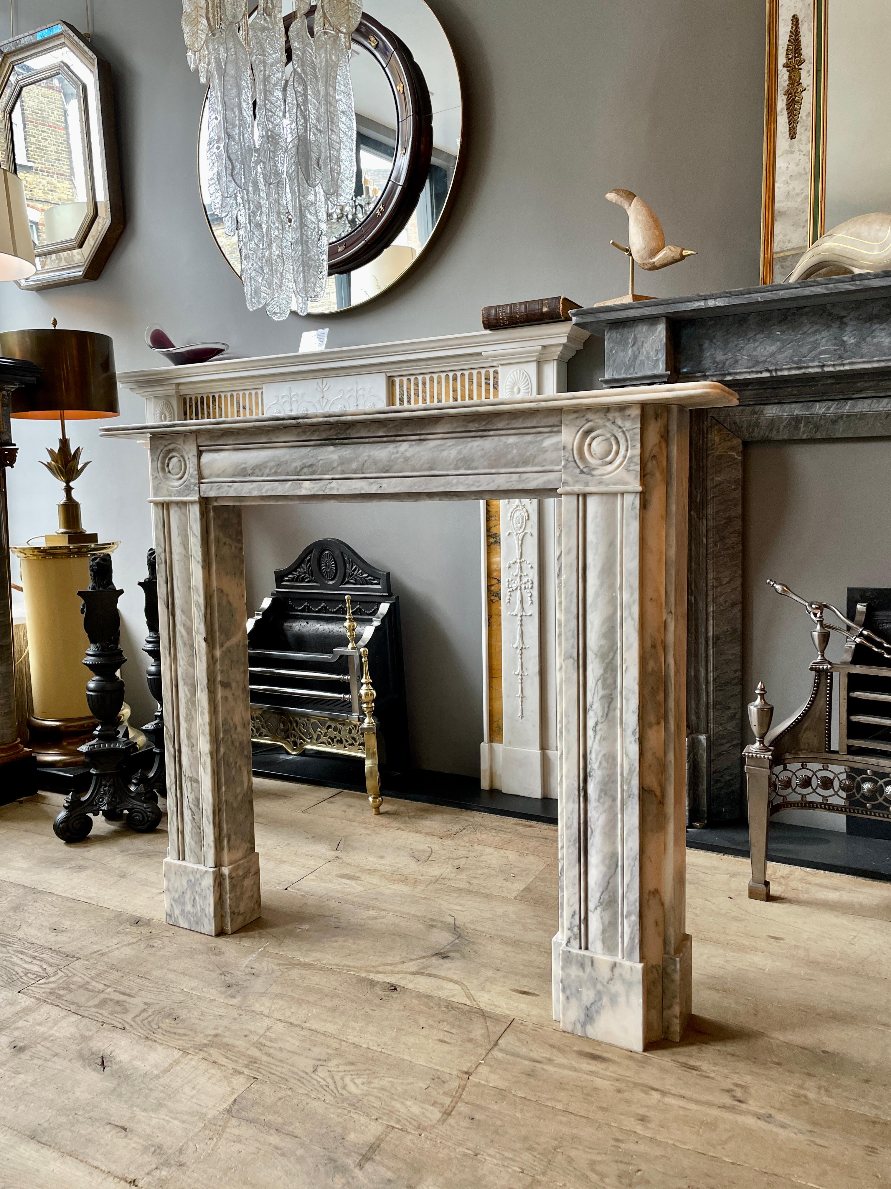 A Regency style fireplace with cushion moulded jambs and front panels on square foot blocks and surmounted by roundel carved corner blocks. Conforming cushion moulded frieze with a simple reeded shelf. Made in Italian Arabescato marble. A reclaimed