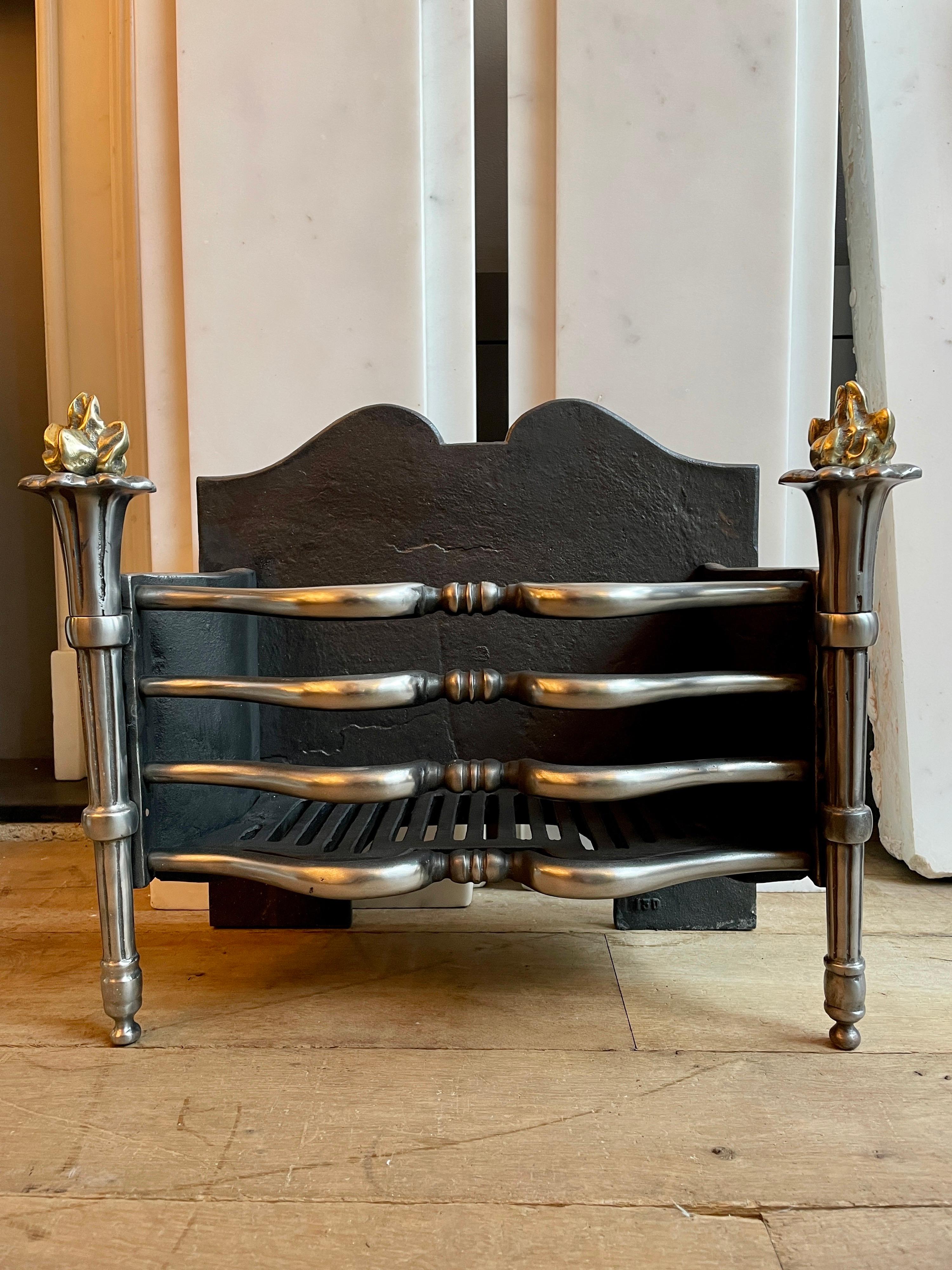 A polished wrought iron fire grate in the regency manner, with 4 shaped bars and torchere supports with brass flame finials. English, early 20th century.