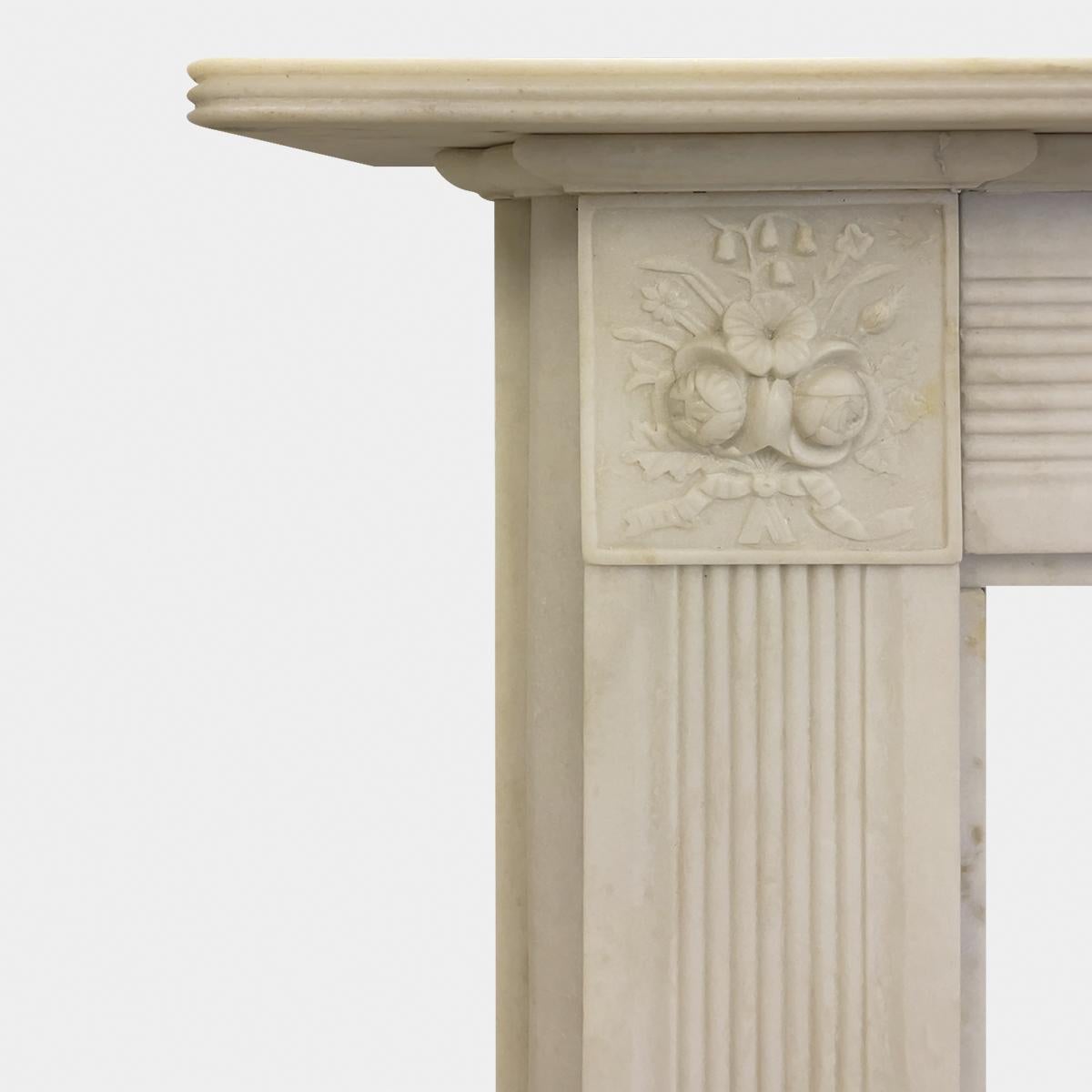 A reclaimed Regency style fireplace in lightly distressed or antiqued white Aegean marble. With reeded panels to the jambs surmounted by carved foliate corner blocks. The frieze also reeded beneath a generous reeded shelf. Carved well and in good