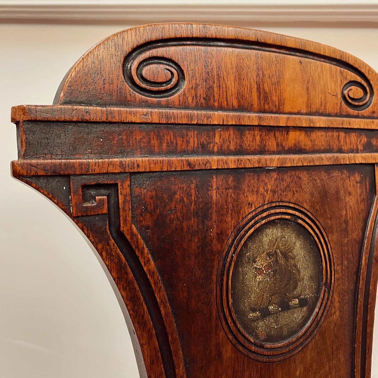 An elegant pair of regency mahogany hall chairs, the waisted panelled back with carved stretched swirls to top rail above Soanian squashed Greek key borders flanking a recessed and reeded roundel with finely painted heraldic crest depicting a